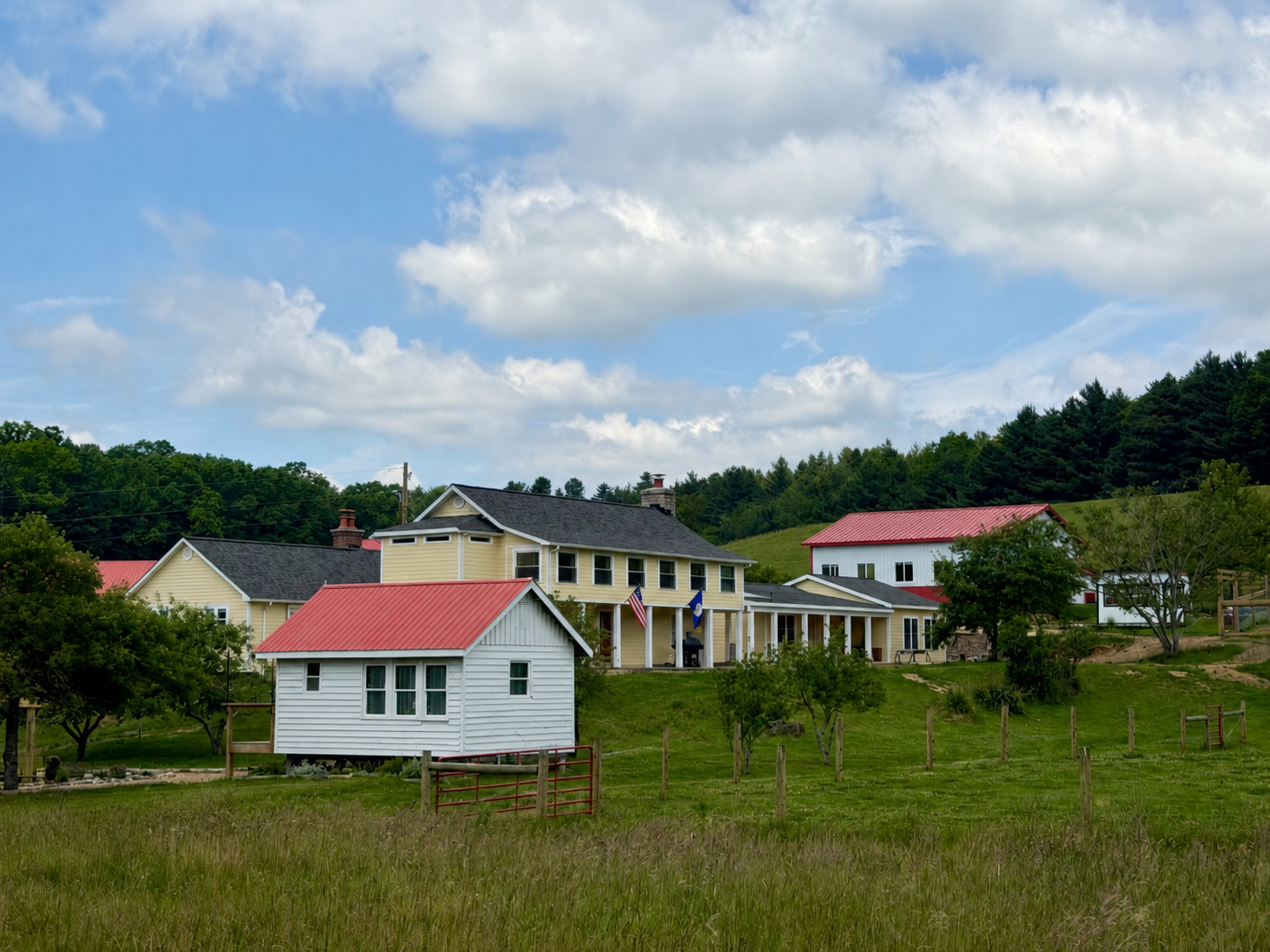 A scenic view of a yellow house with white trim, surrounded by smaller white and red outbuildings, set against a lush green hillside with trees, next to a reflective body of water.