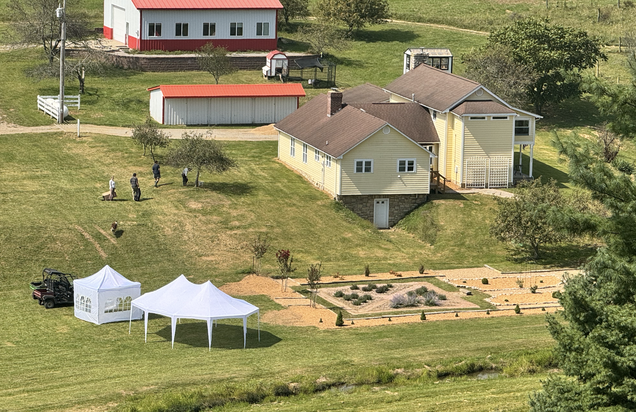 A yellow house with a brown roof, a front porch, and a stone foundation, situated on a grassy hillside. There are two tents and a golf cart nearby, with a garden and trees surrounding the area. Four people are walking near the house.