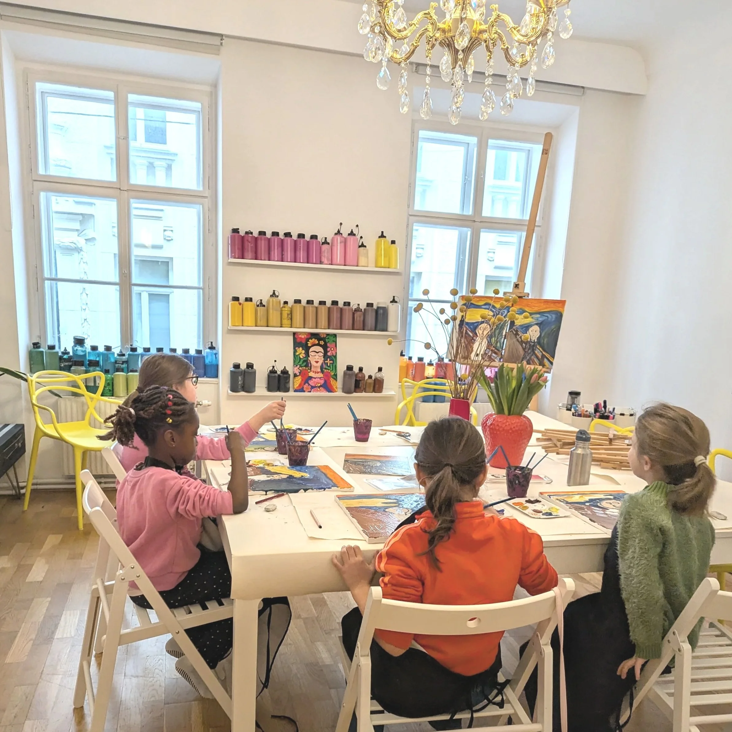 Four children painting at a table in an art classroom with colorful shelves, a chandelier, and Vincent van Gogh's 'The Starry Night' painting.