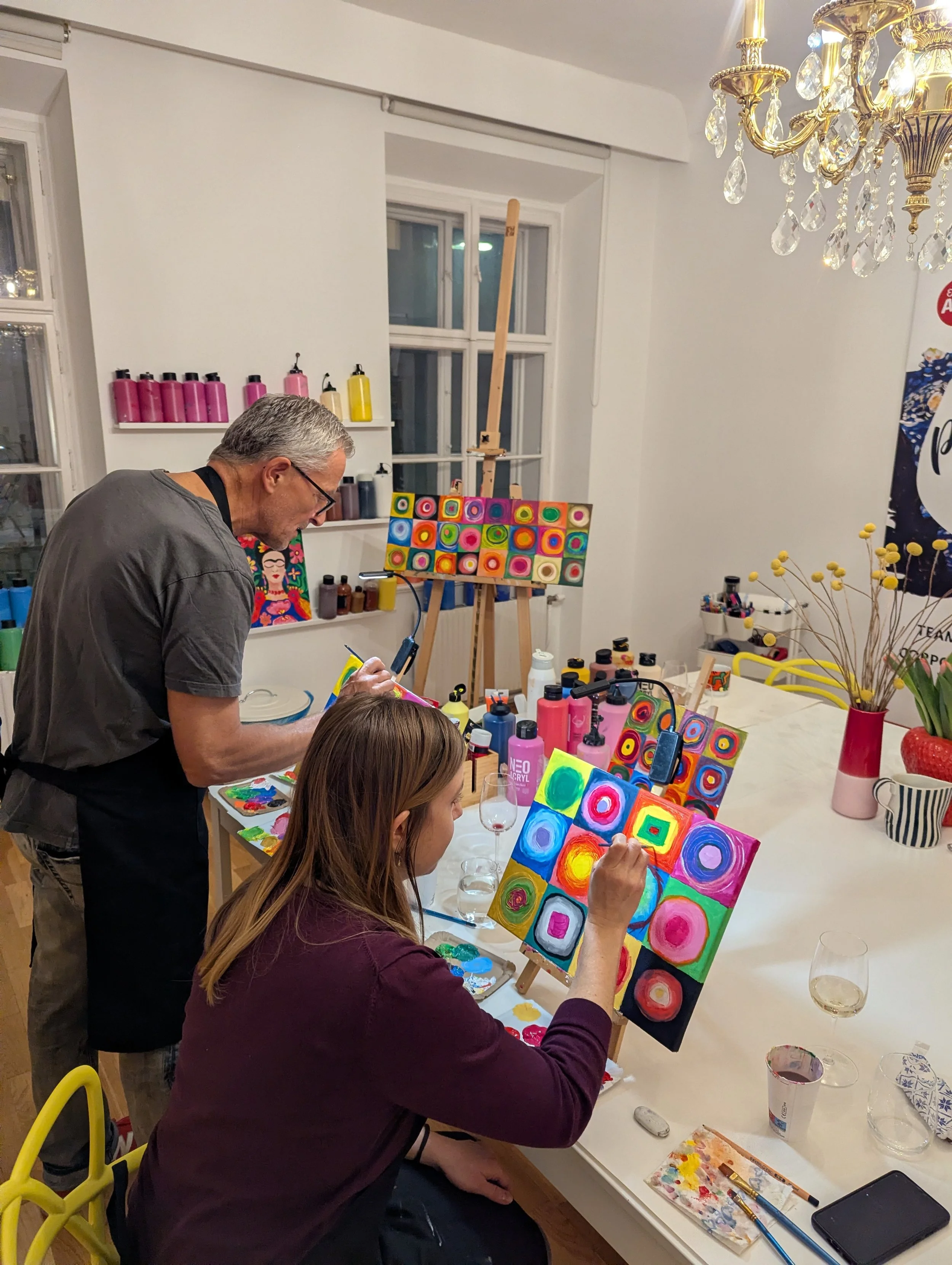 Two women painting colorful abstract artwork on small canvases while a man observes in a cozy, well-lit art studio with colorful supplies and finished paintings on shelves.