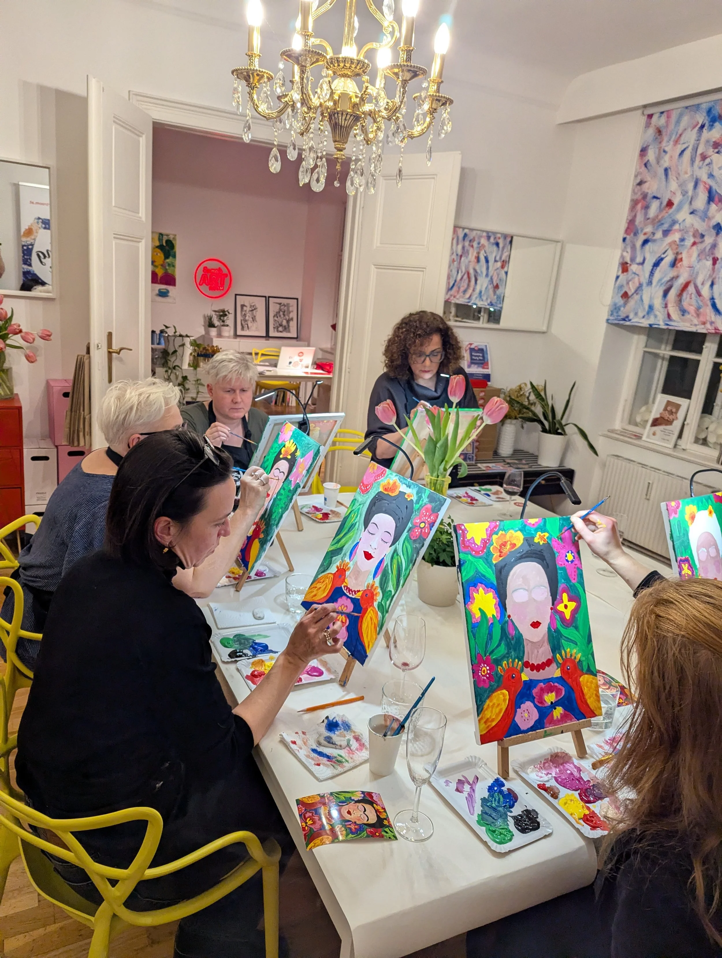 A group of women participating in a painting class around a table, with colorful portraits of a woman with a bun and flowers, using brushes and paint palettes, in a well-lit room with a chandelier and abstract curtains.