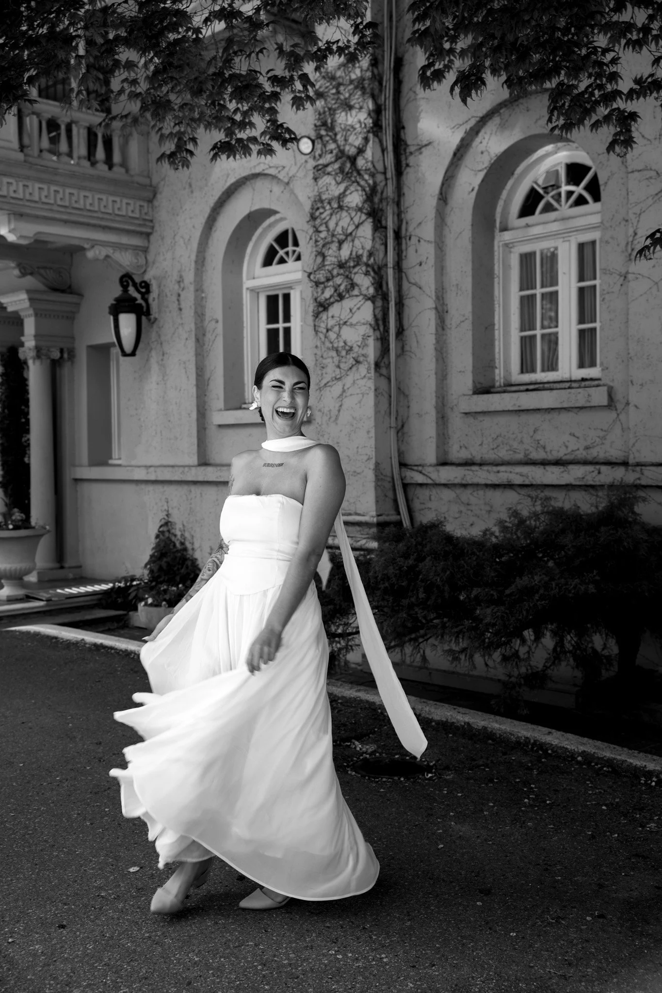 A woman in a white strapless dress joyfully spinning outdoors in front of a building with arched windows and ivy-covered walls, captured in black and white.