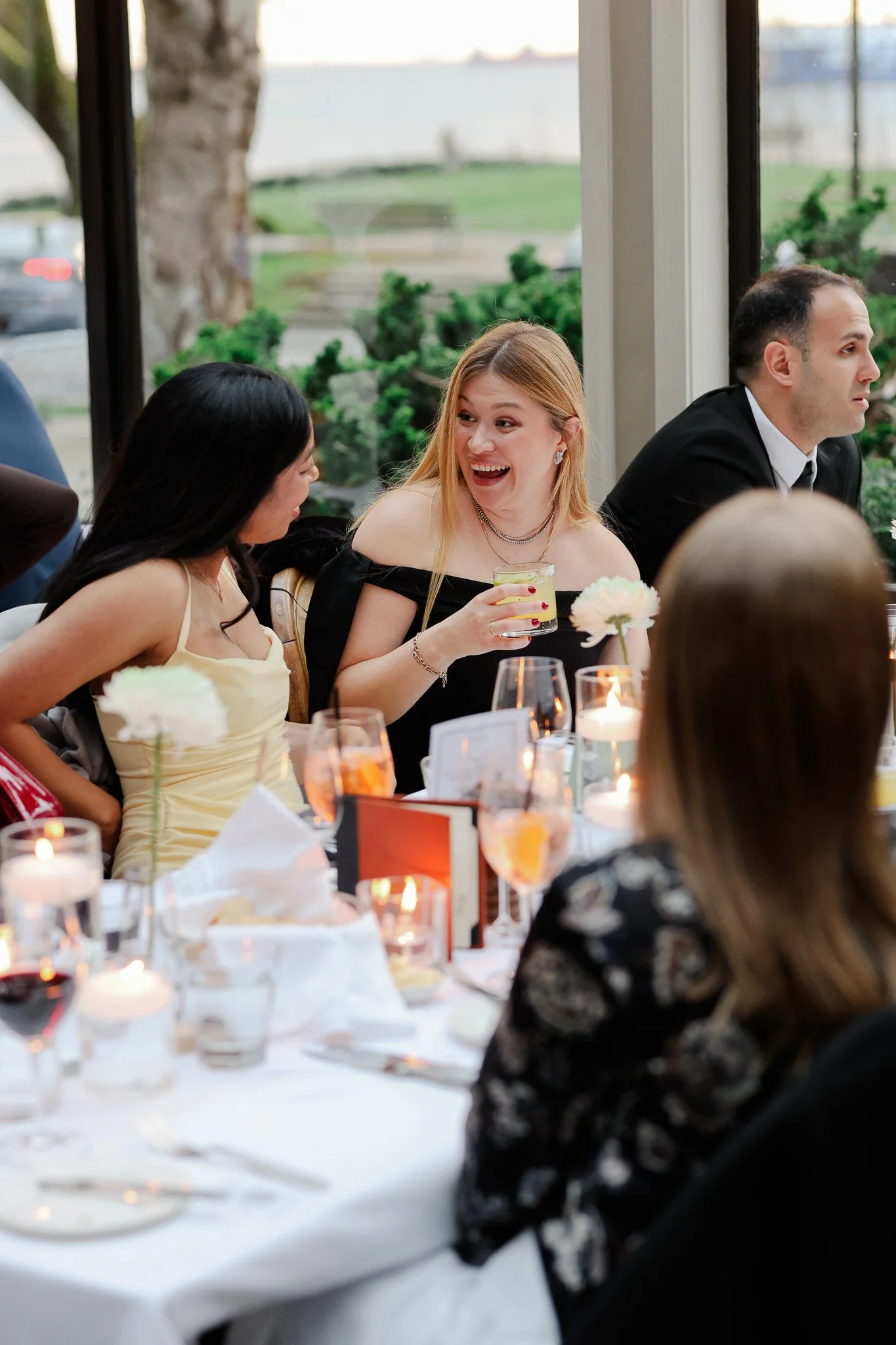 People sitting at a banquet table with flowers, candles, and drinks, enjoying a social event in a room with large windows showing an outdoor landscape.