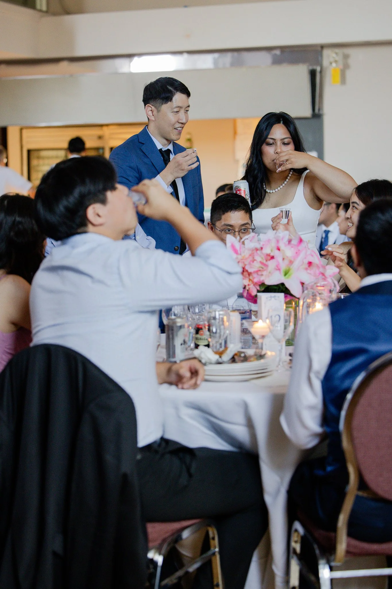 Group of people at a formal event, some drinking and smiling, with a woman in a white dress and a man in a blue suit.