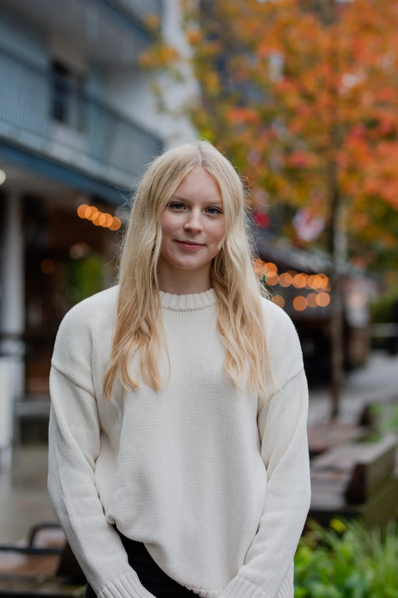 A young woman with blonde hair and fair skin stands outdoors with autumn trees and a building in the background. She is wearing a cream-colored sweater and smiling gently.