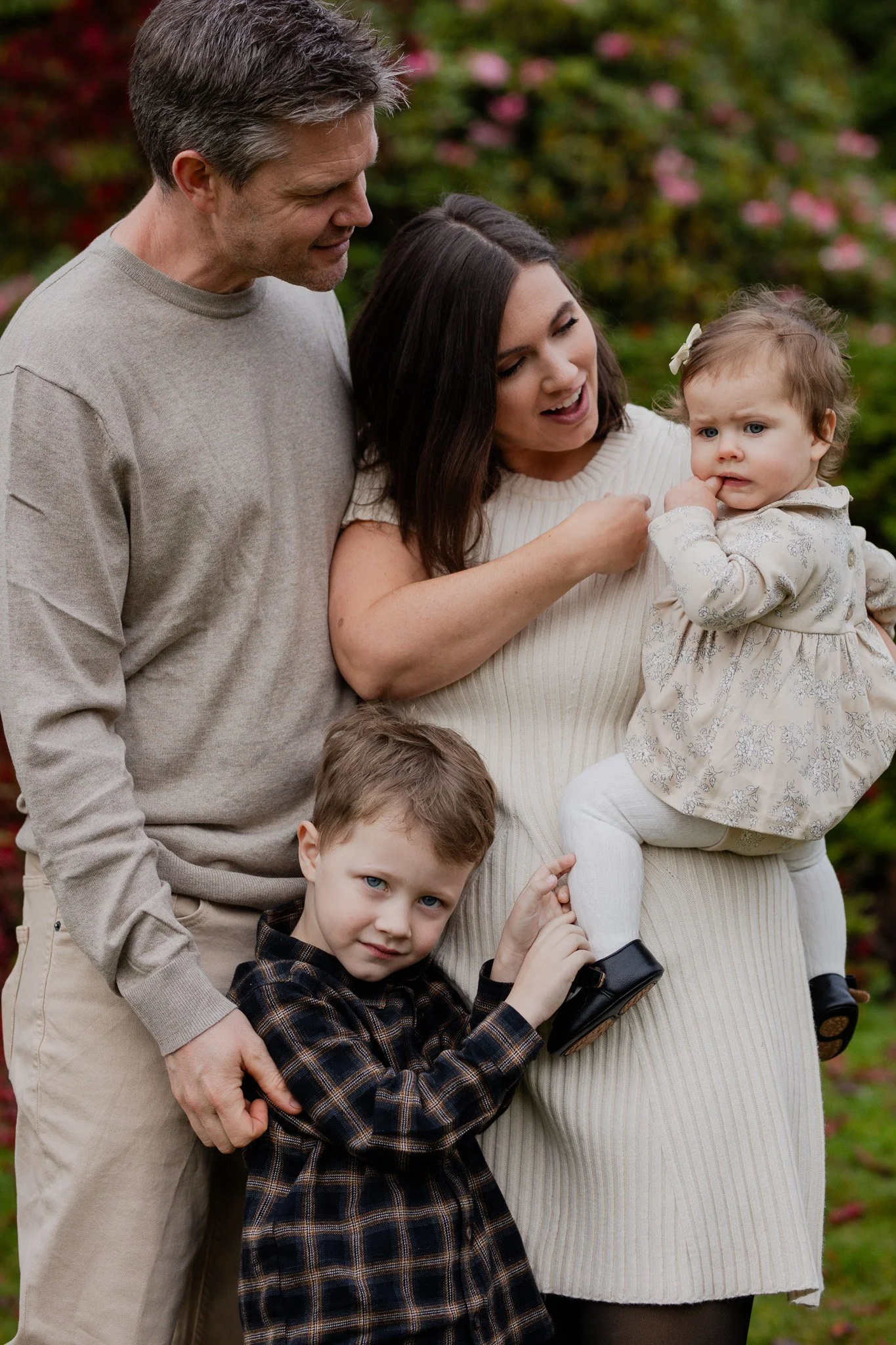 Family of four standing outdoors with pink flowers and green trees in the background, smiling and interacting with each other.