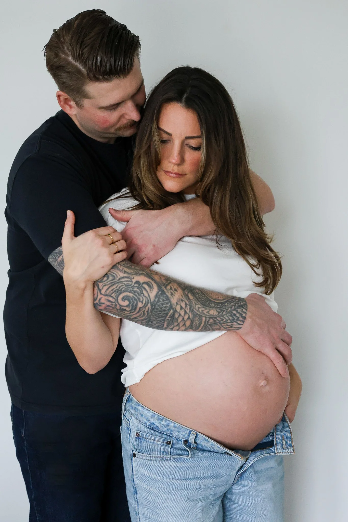A man embracing a pregnant woman, holding her belly with one hand, her eyes closed, against a plain white background.