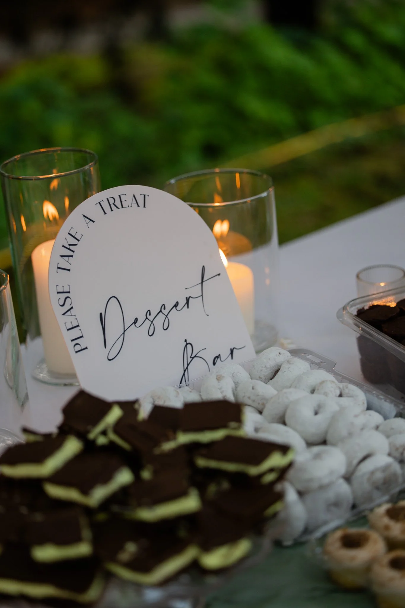 A dessert table with cookies, chocolates, and desserts, with a sign that reads "Please Take A Treat" and candles in the background.