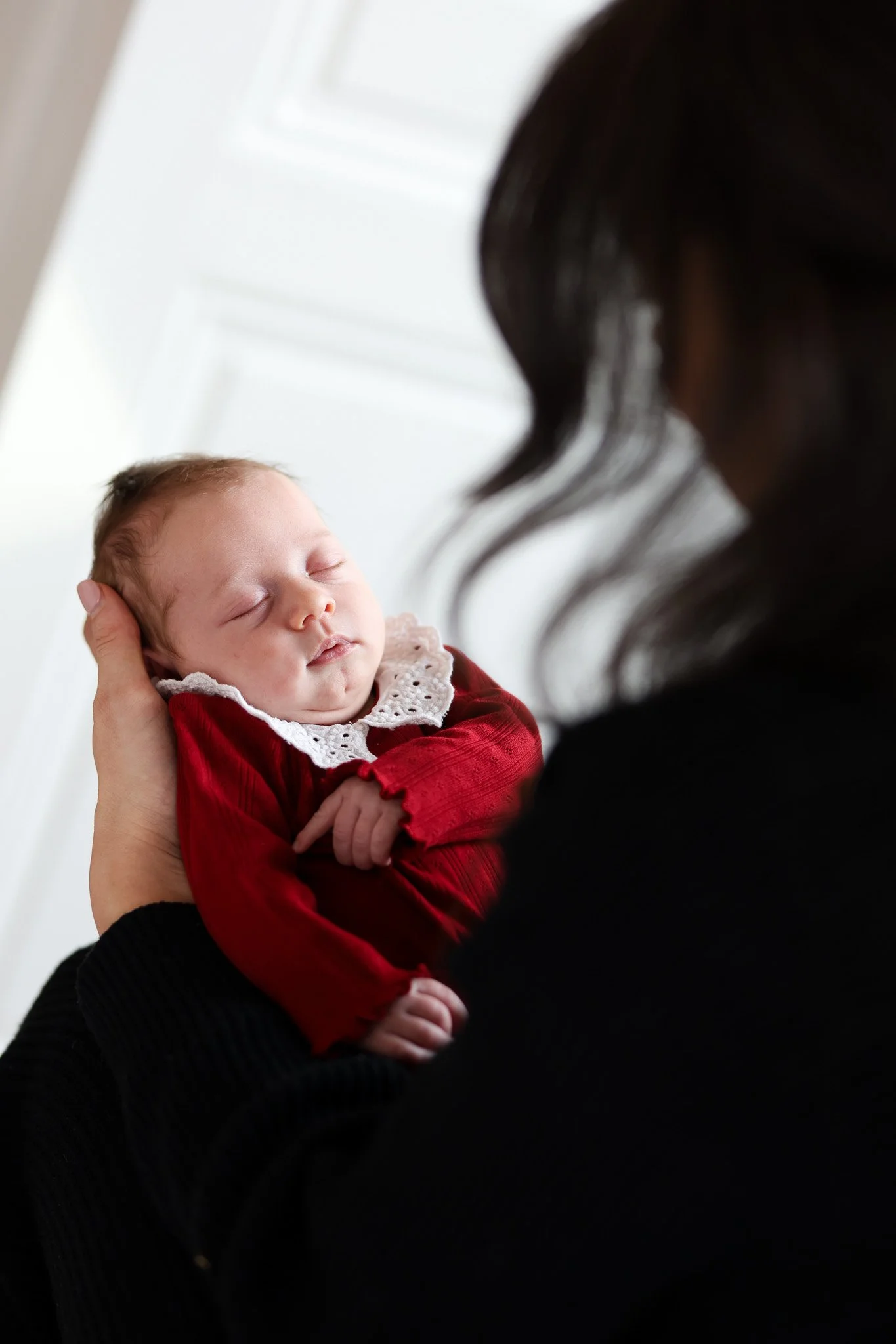 A woman holds a sleeping baby dressed in red with a white lace collar, against a white background.
