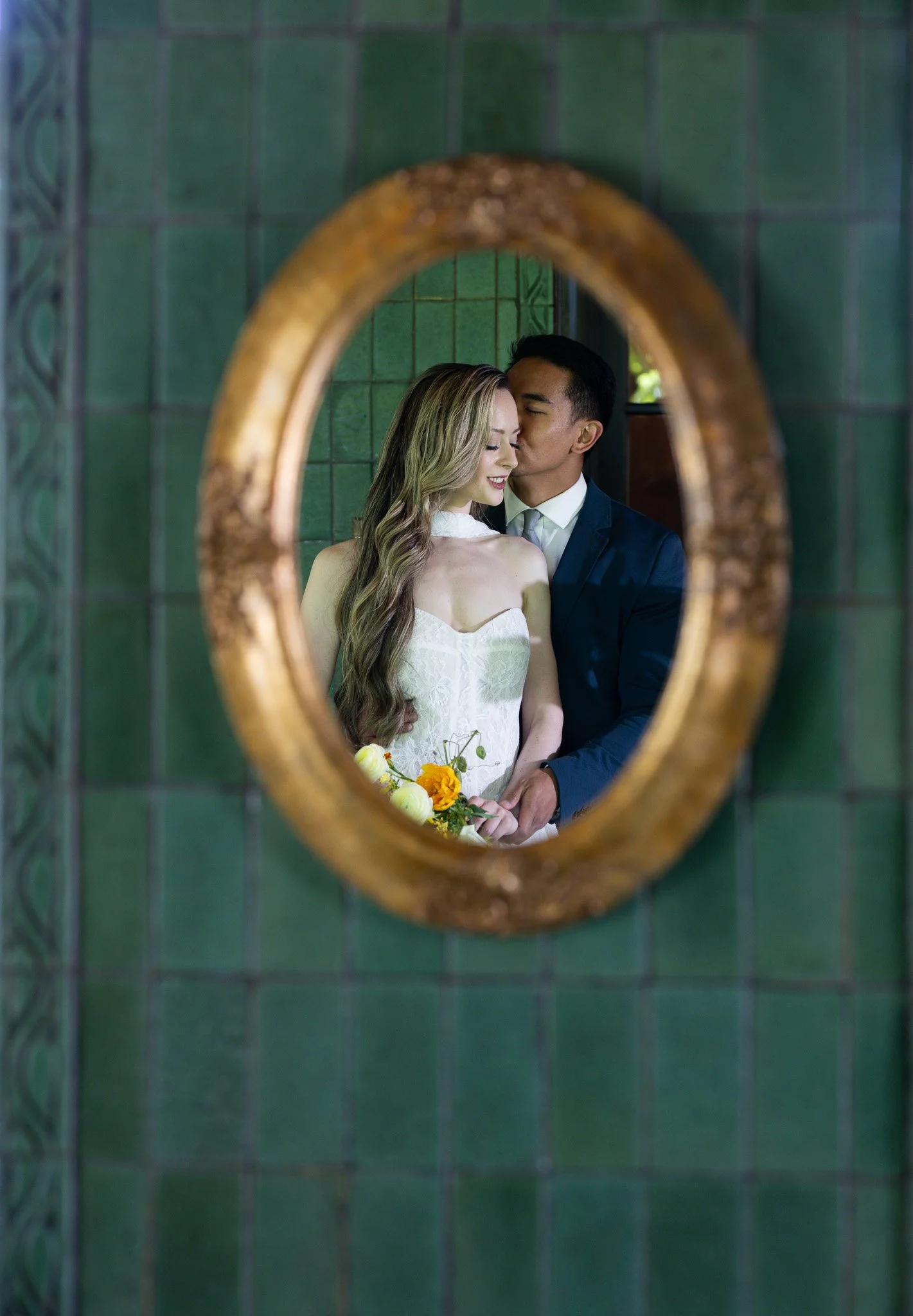 Reflection of a bride and groom in an oval mirror with a gold frame, green tiled wall background, bride holding a bouquet of yellow, white, and orange flowers, groom kissing her cheek.