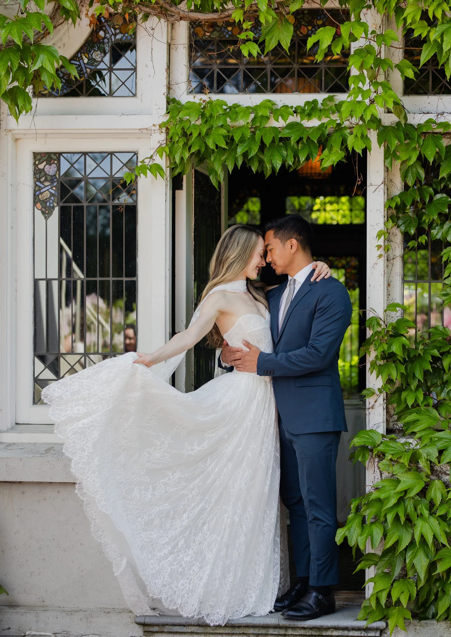 A couple dressed in wedding attire, embracing and touching foreheads in front of a white building with green ivy and stained glass windows.
