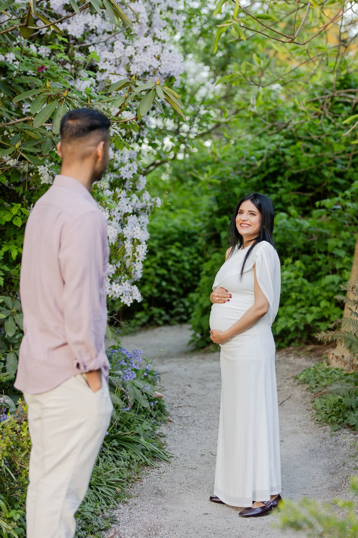 A woman in a white dress smiling at a man in a pink shirt and cream pants in a garden with green foliage and blooming flowers.