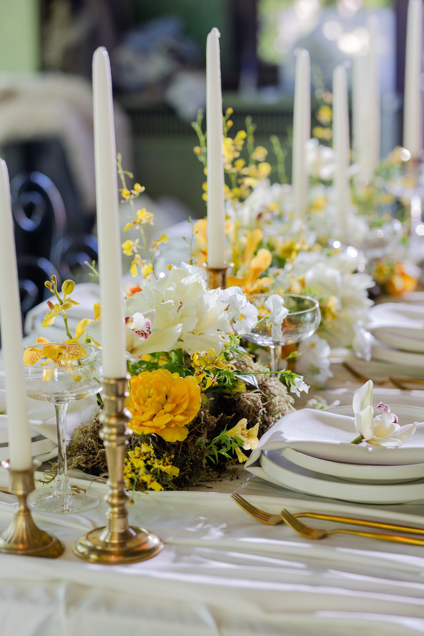 Elegant dining table with white candles, floral centerpiece with white and yellow flowers, gold utensils, and white napkins on a white tablecloth.