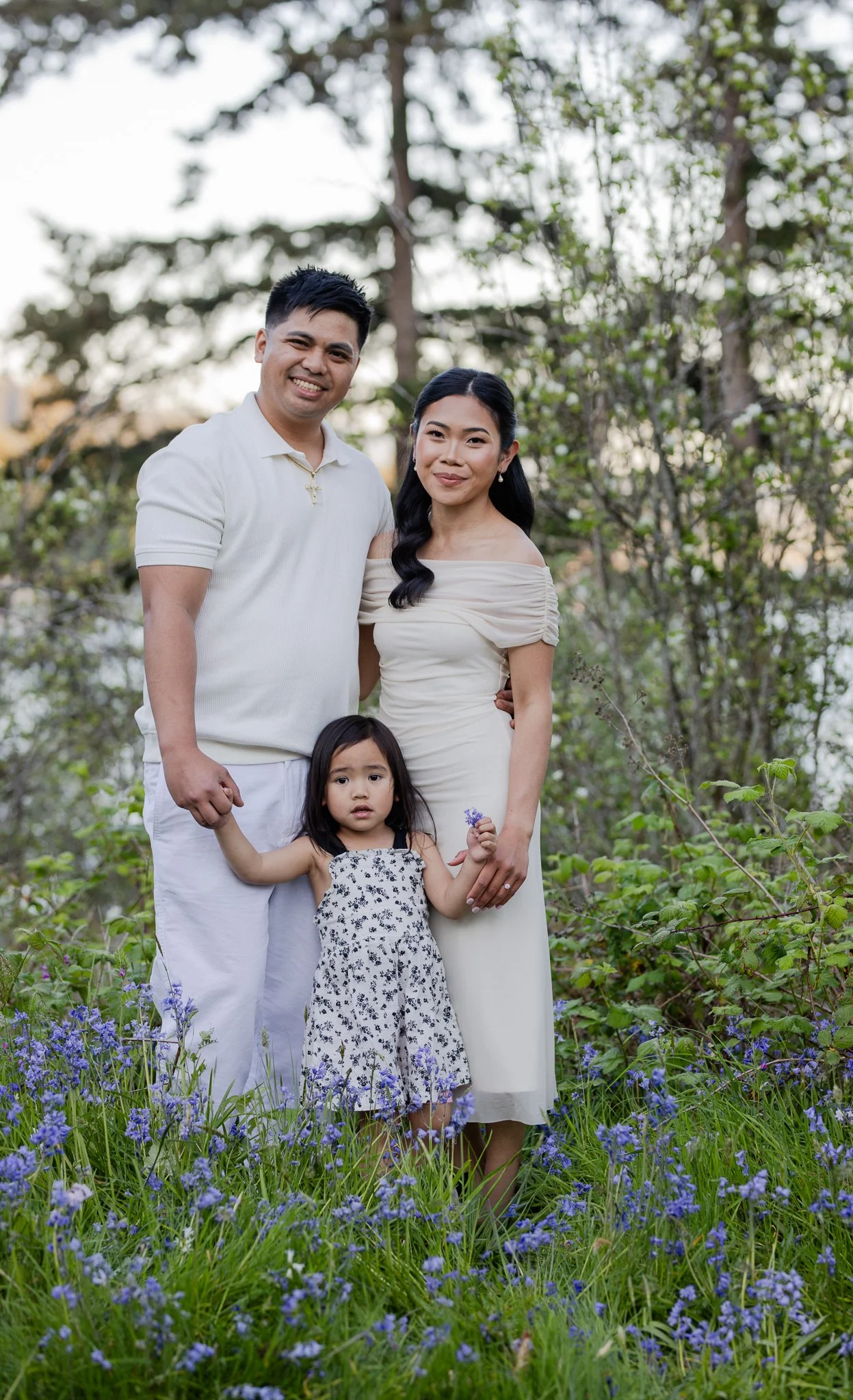 A family of three, including a man, woman, and young girl, standing outdoors in a garden with purple flowers and trees in the background, smiling and posing for the photo.