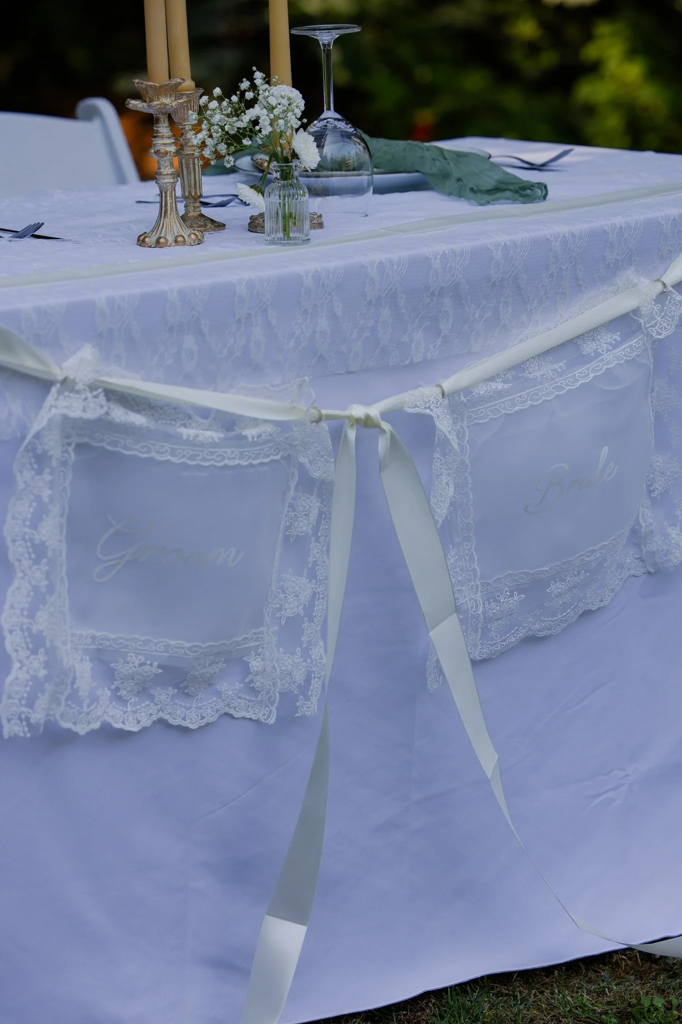 A close-up of a decorated outdoor table with a white lace tablecloth, floral centerpiece, glassware, and candles, set for an event.
