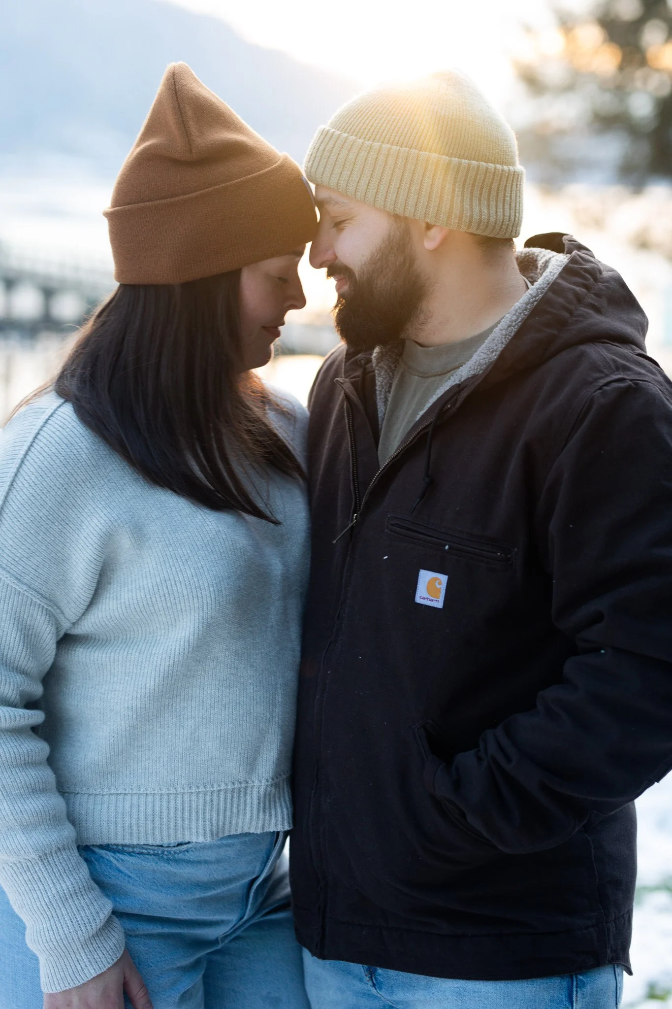 A couple standing close together with foreheads touching outdoors during sunset, wearing casual winter clothing and beanies.