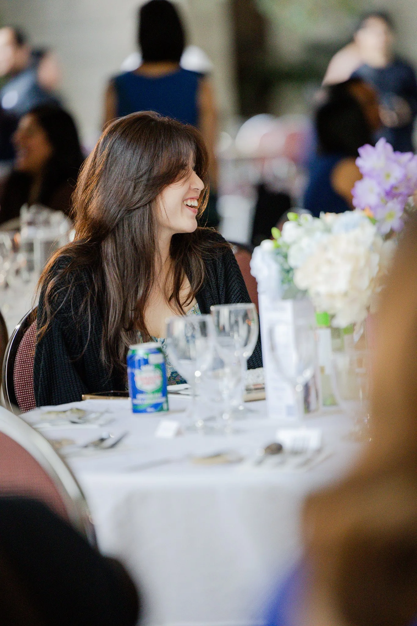 A woman with brown hair smiling at a formal event, sitting at a table with flower arrangements, glassware, and a can of sparkling water.