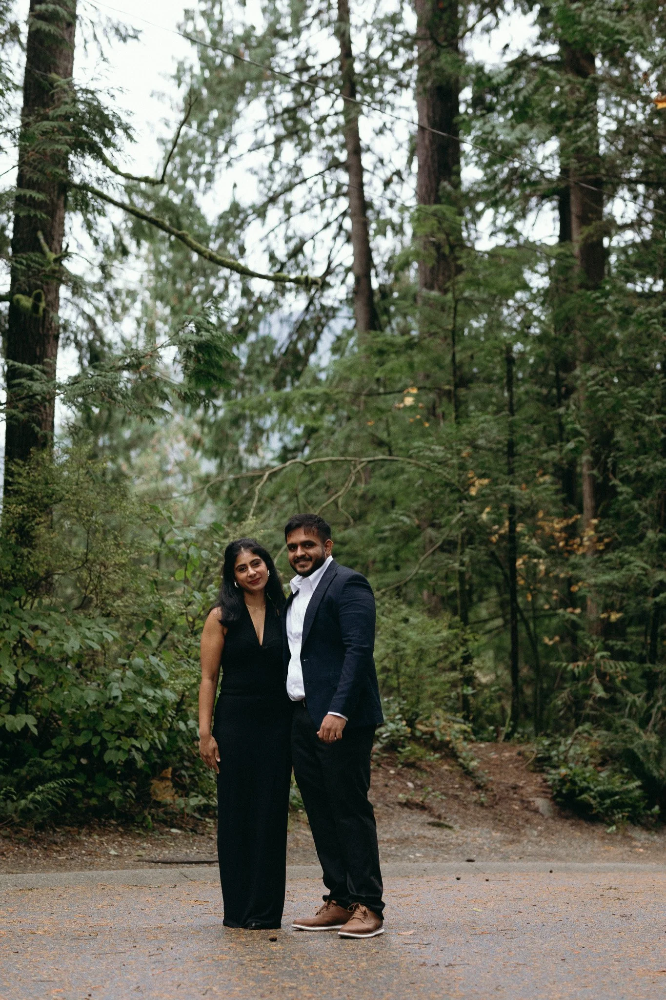A man and a woman in formal attire standing on a forest path, surrounded by tall trees and green foliage.