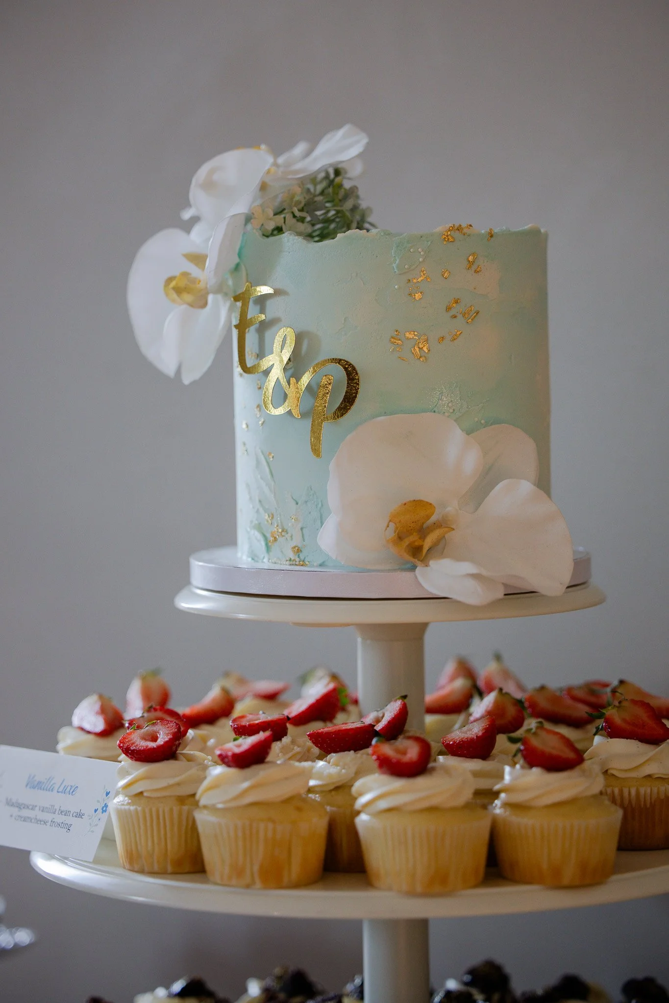 Tiered dessert display with a front row of cupcakes topped with strawberries and cream, a middle row of vanilla bean cake with cream frosting and strawberries, and an upper tier with a pastel-colored cake decorated with flowers, gold accents, and the