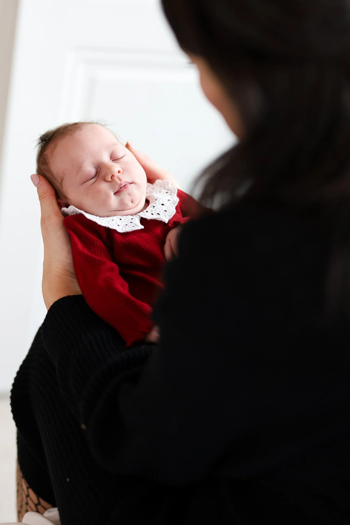 A woman holding a sleeping baby dressed in a red outfit with a lace collar. The woman is partially visible with dark hair, and the background is white.