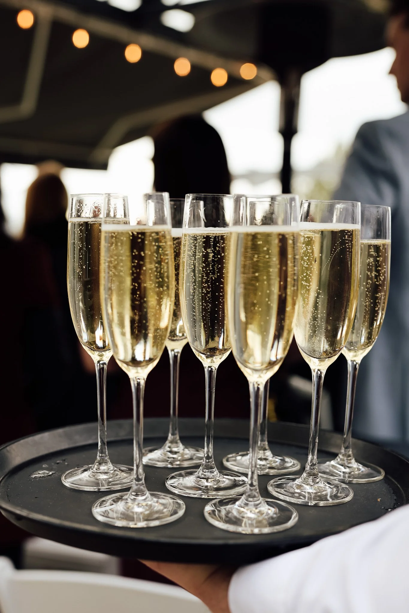 A tray of 10 champagne flutes filled with sparkling champagne, with bubbles rising to the surface, being carried by a person wearing a white shirt at an event or celebration.