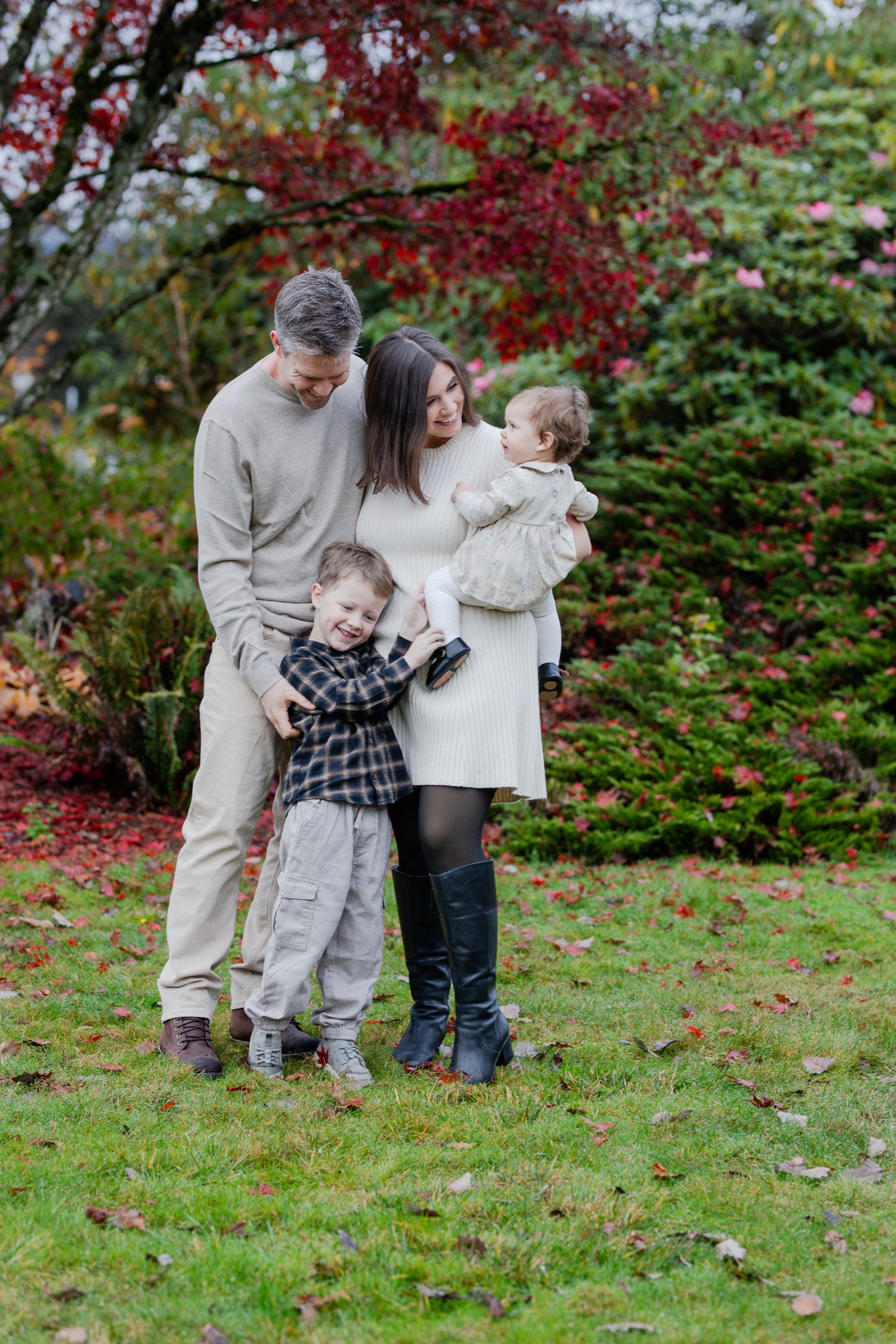 Family of four outdoors in a garden during fall, smiling, with colorful trees and bushes in background.