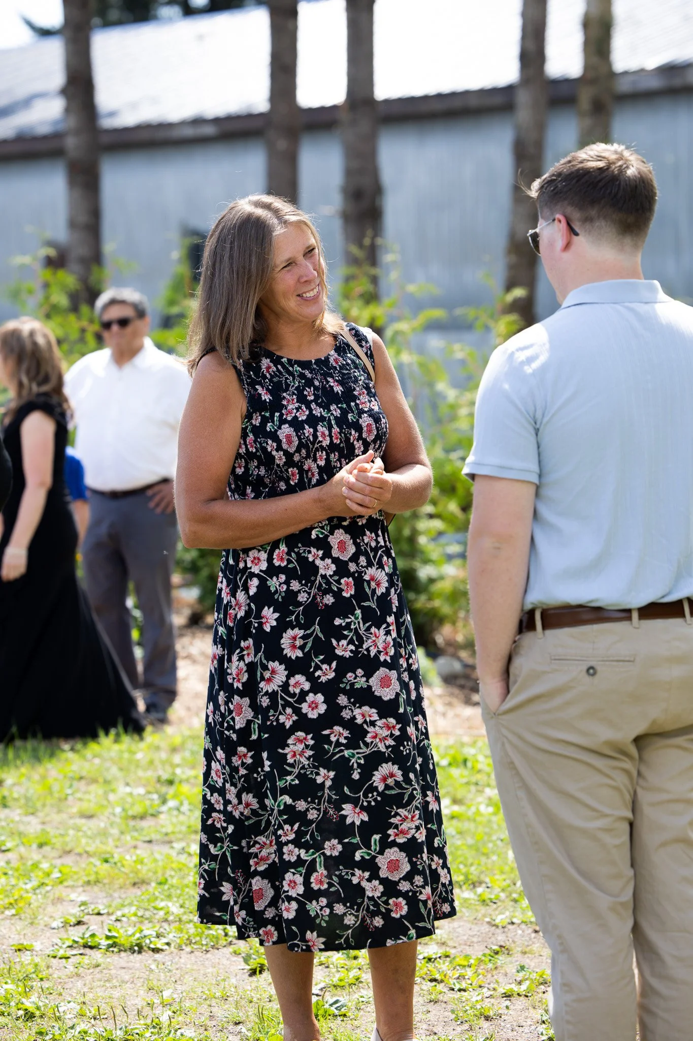 A woman in a black floral dress smiling and talking to a man in sunglasses and light-colored clothing outdoors, with other people and trees in the background.