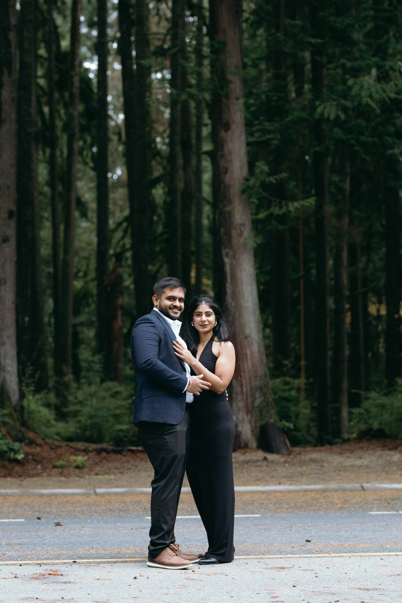 A couple in formal attire standing on a road in front of a forest.