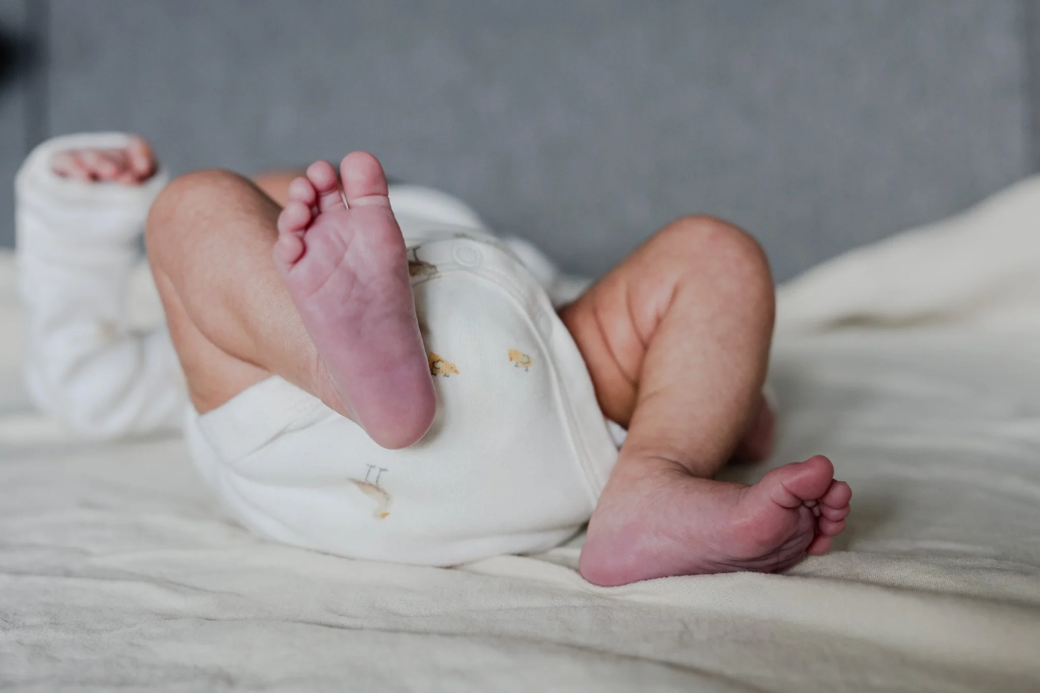 Close-up of a baby's foot and an adult's hand resting on a white surface. The baby is wearing a white onesie with small yellow and brown patterns. The background is blurred and gray.