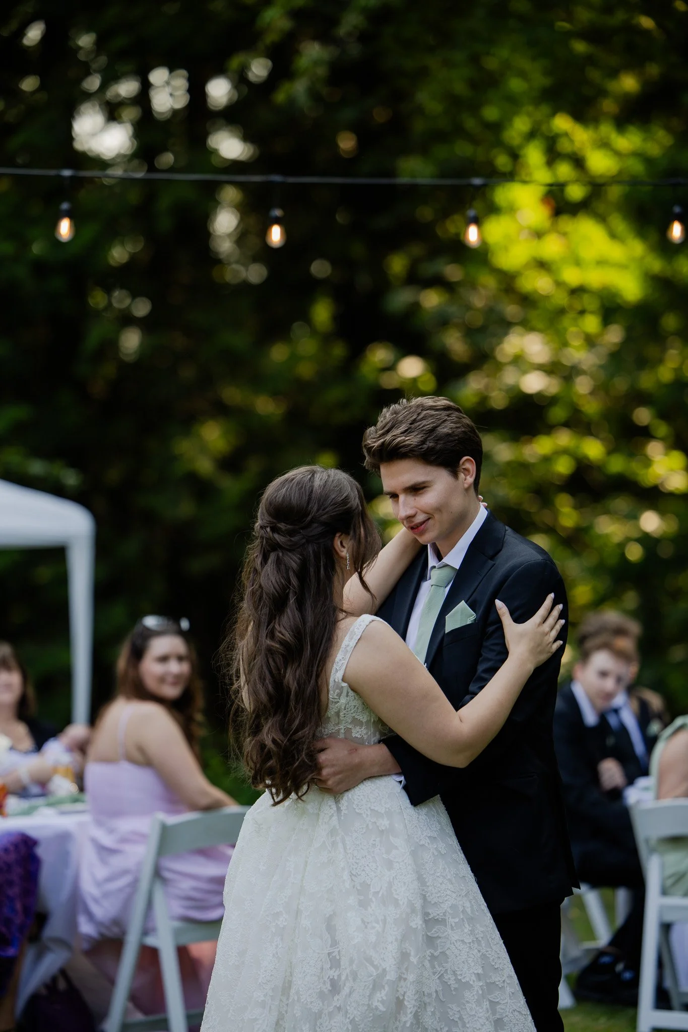 A bride and groom are dancing together at an outdoor wedding reception surrounded by guests. The bride has long dark hair and is wearing a lace wedding gown. The groom has dark hair and is wearing a dark suit with a tie. They are gazing into each oth