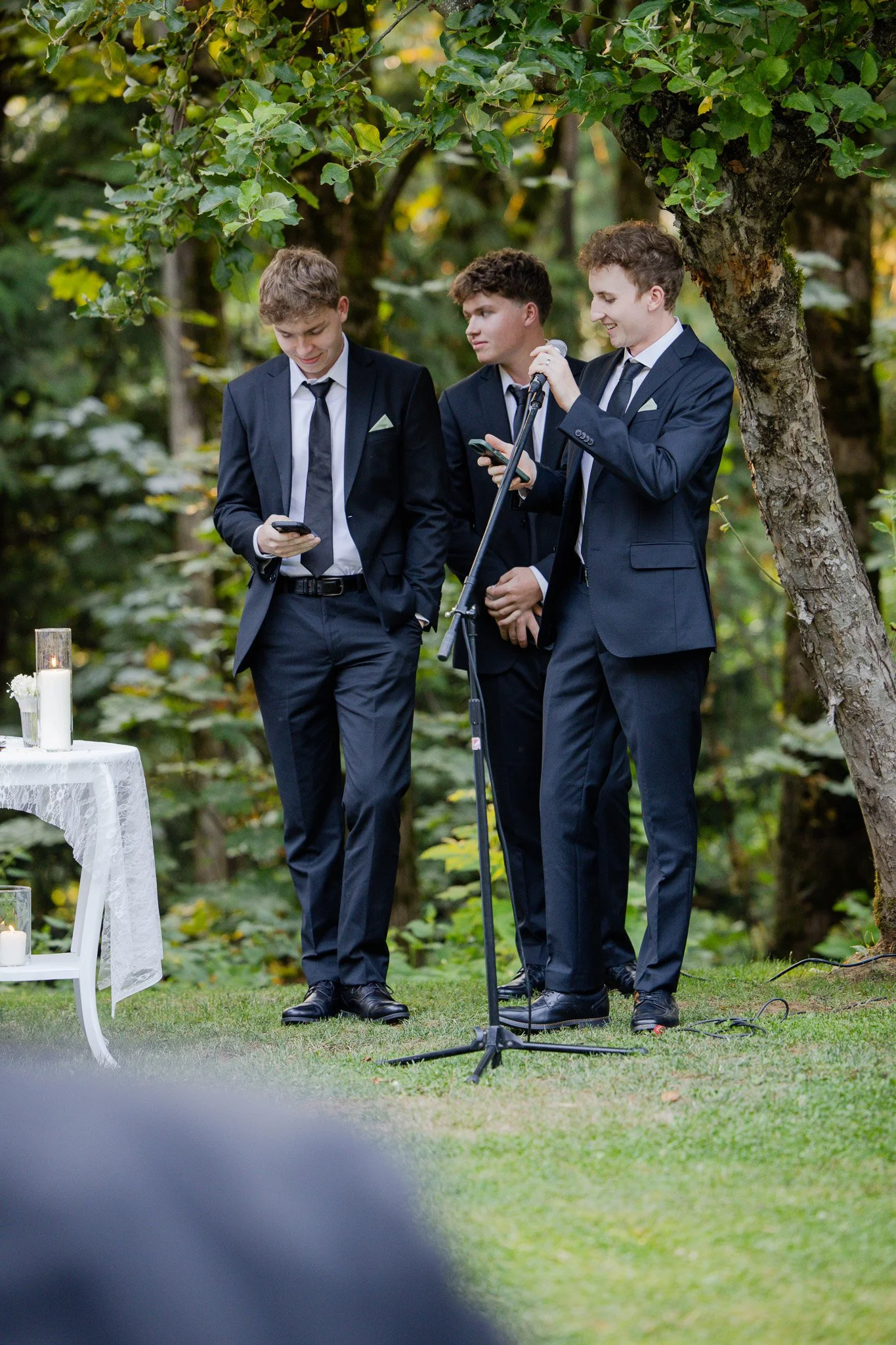 Three young men in formal suits standing outdoors under a tree, with one speaking into a microphone and the others looking at their phones, during a ceremony or event.