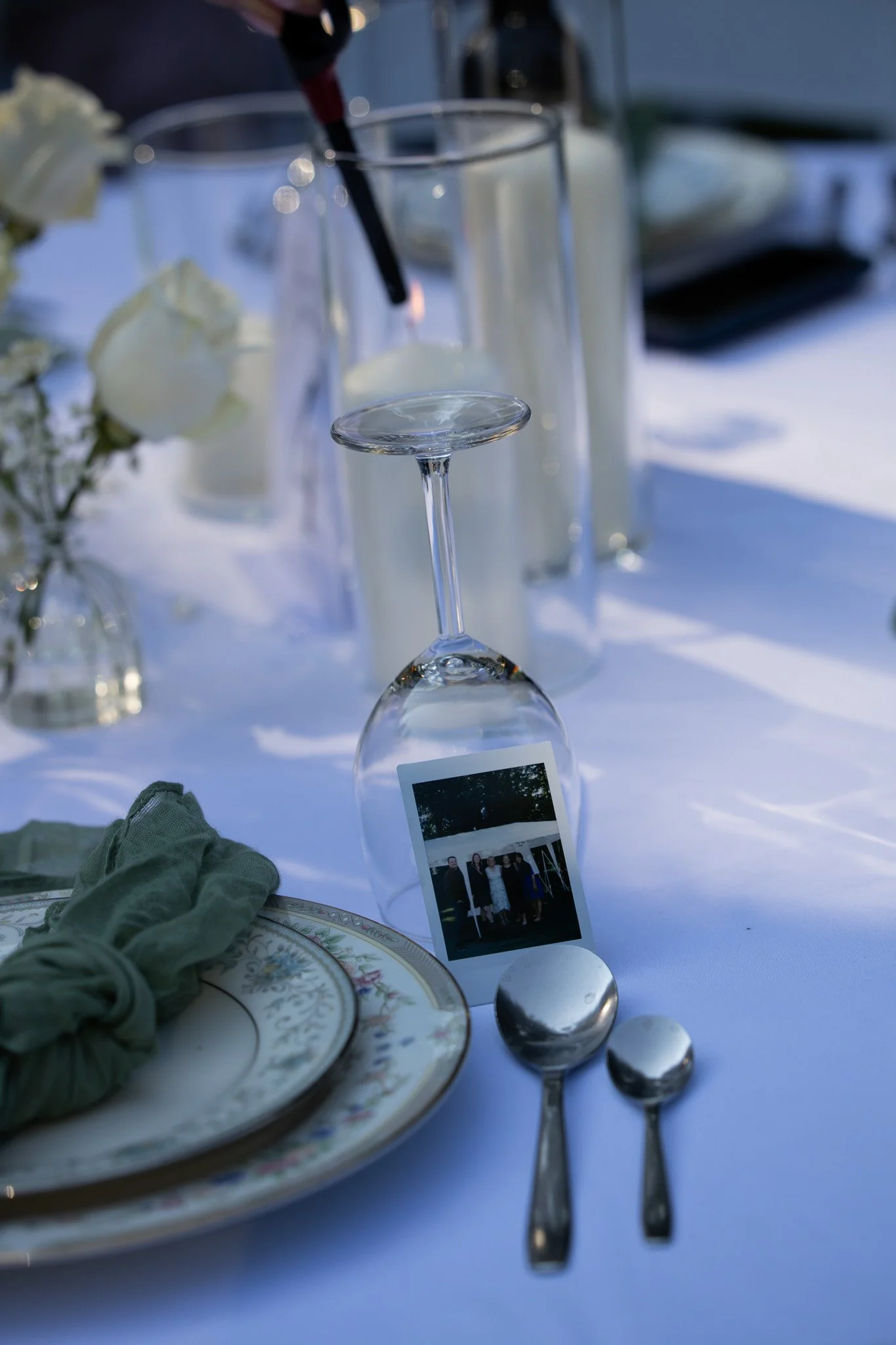 A table set for an outdoor event with a white tablecloth, featuring a champagne flute, silverware, and a small polaroid photograph of four people, with a floral plate and gloves on the table.