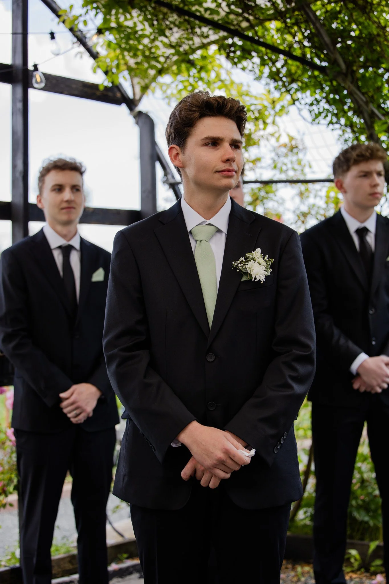 A group of young men dressed in formal black suits and ties, with boutonnieres, attending an outdoor event under a canopy of trees.