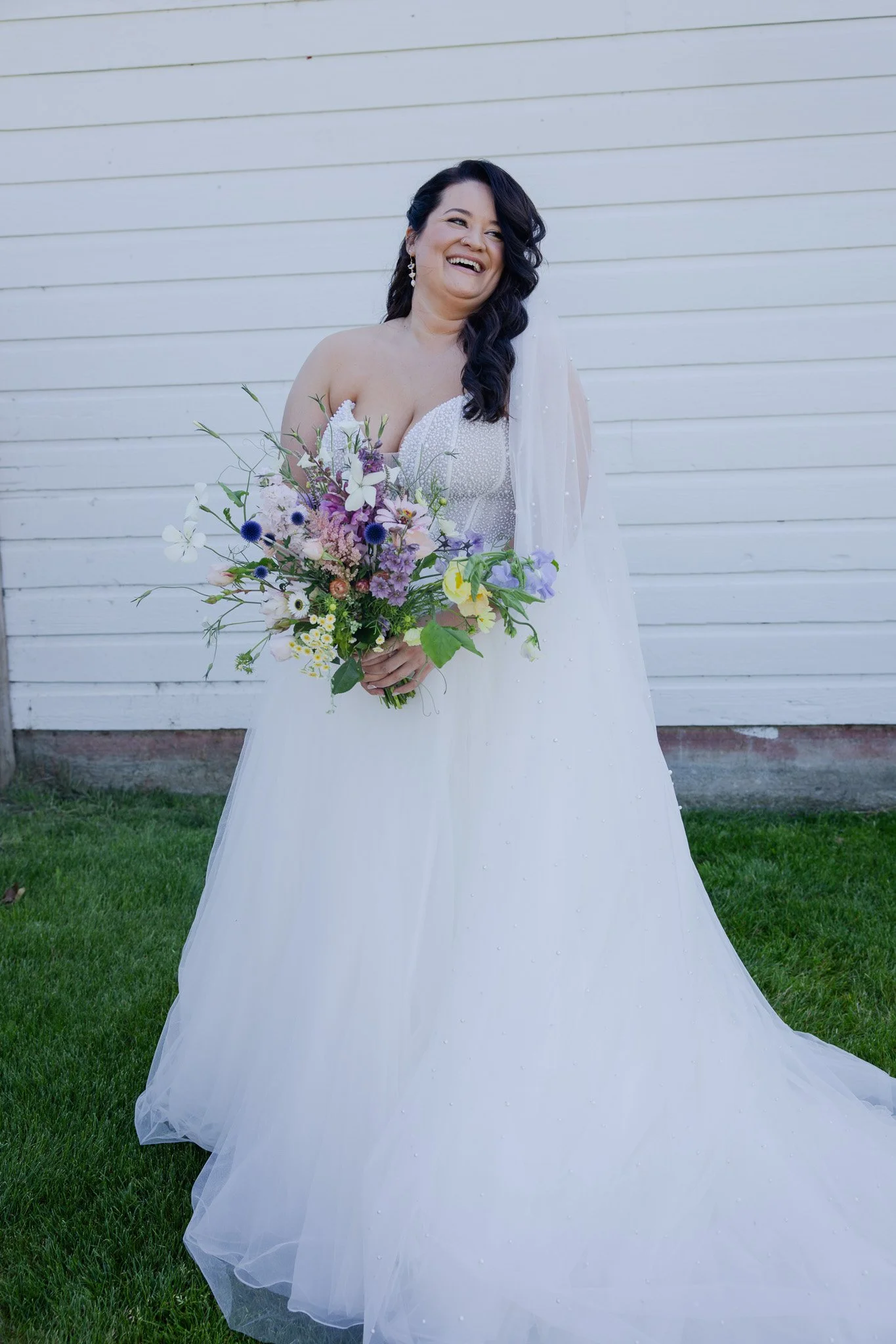 A smiling woman in a wedding dress holding a bouquet of flowers, standing outdoors against a white wooden wall.