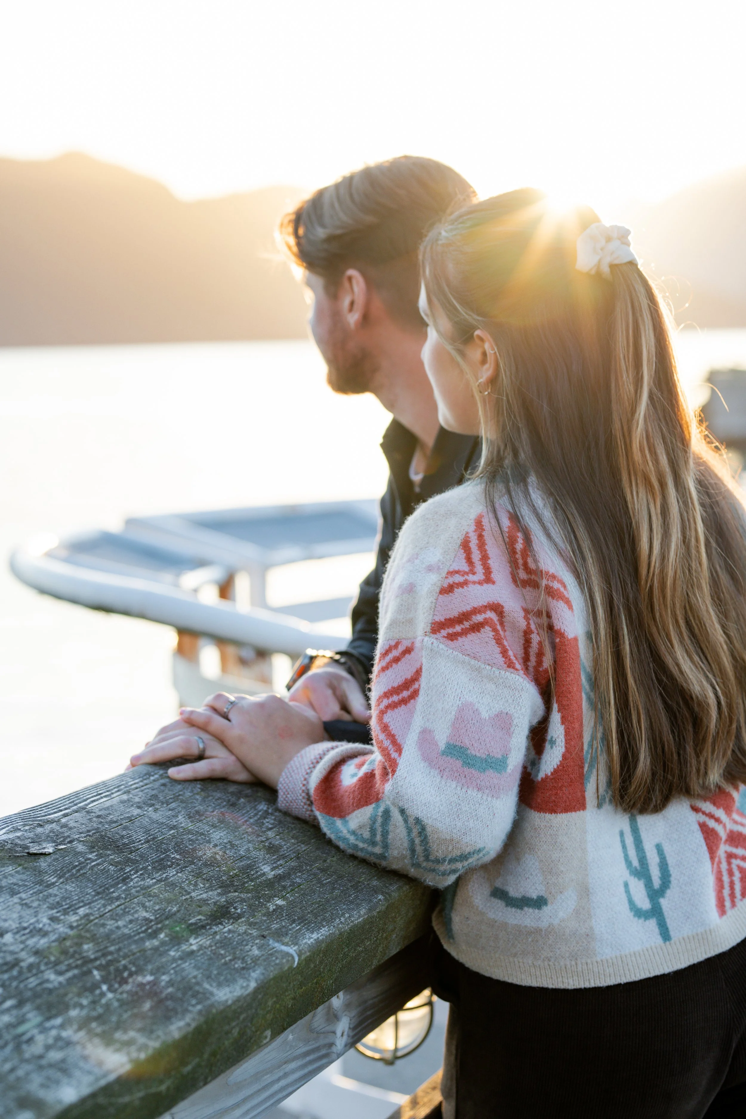 A young man and woman are standing by a wooden railing outdoors near a body of water at sunset, holding hands and looking into the distance.