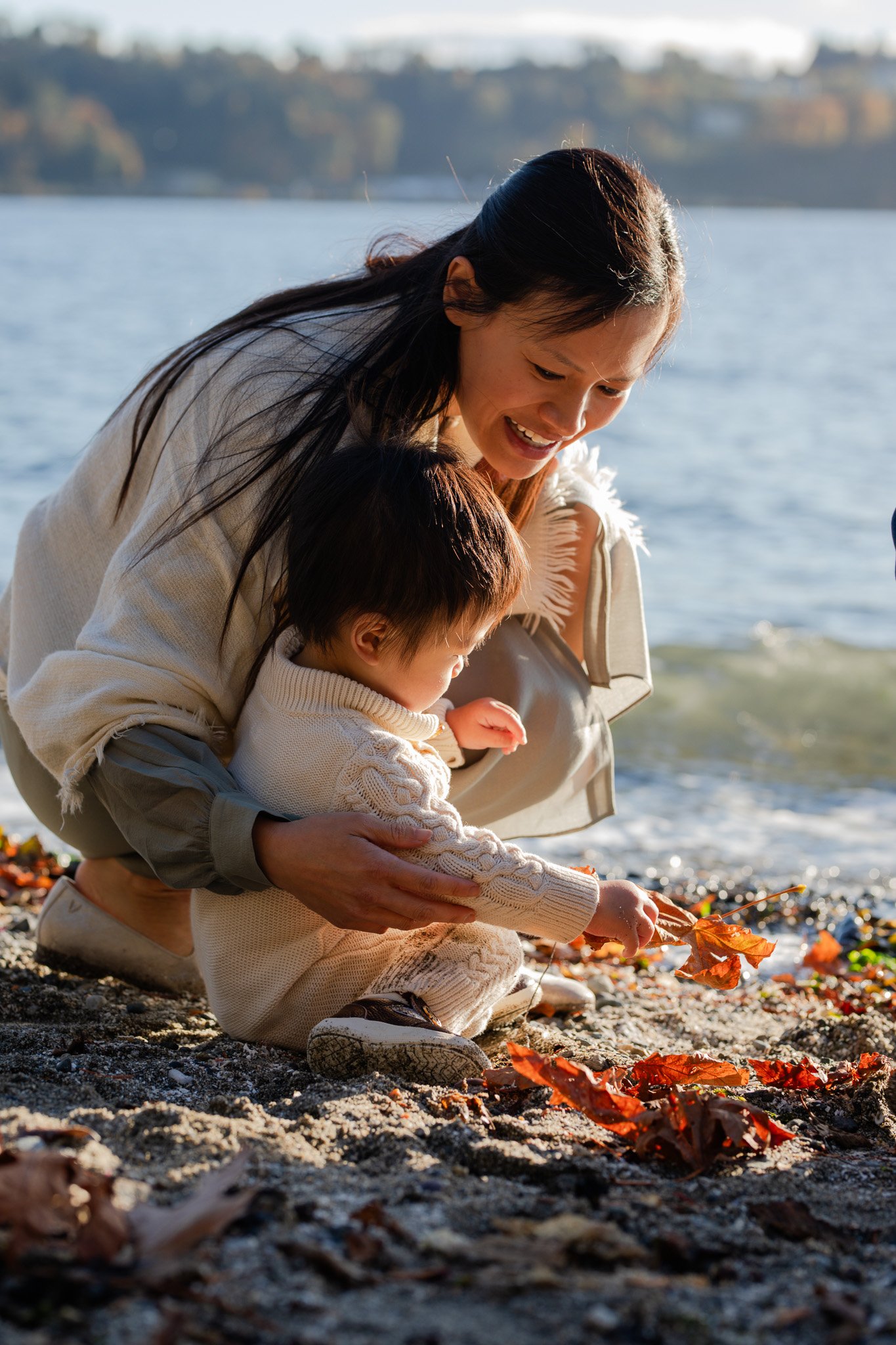 A woman and a young child playing with fallen leaves on a beach by a lake during autumn, smiling and enjoying the outdoor scenery.