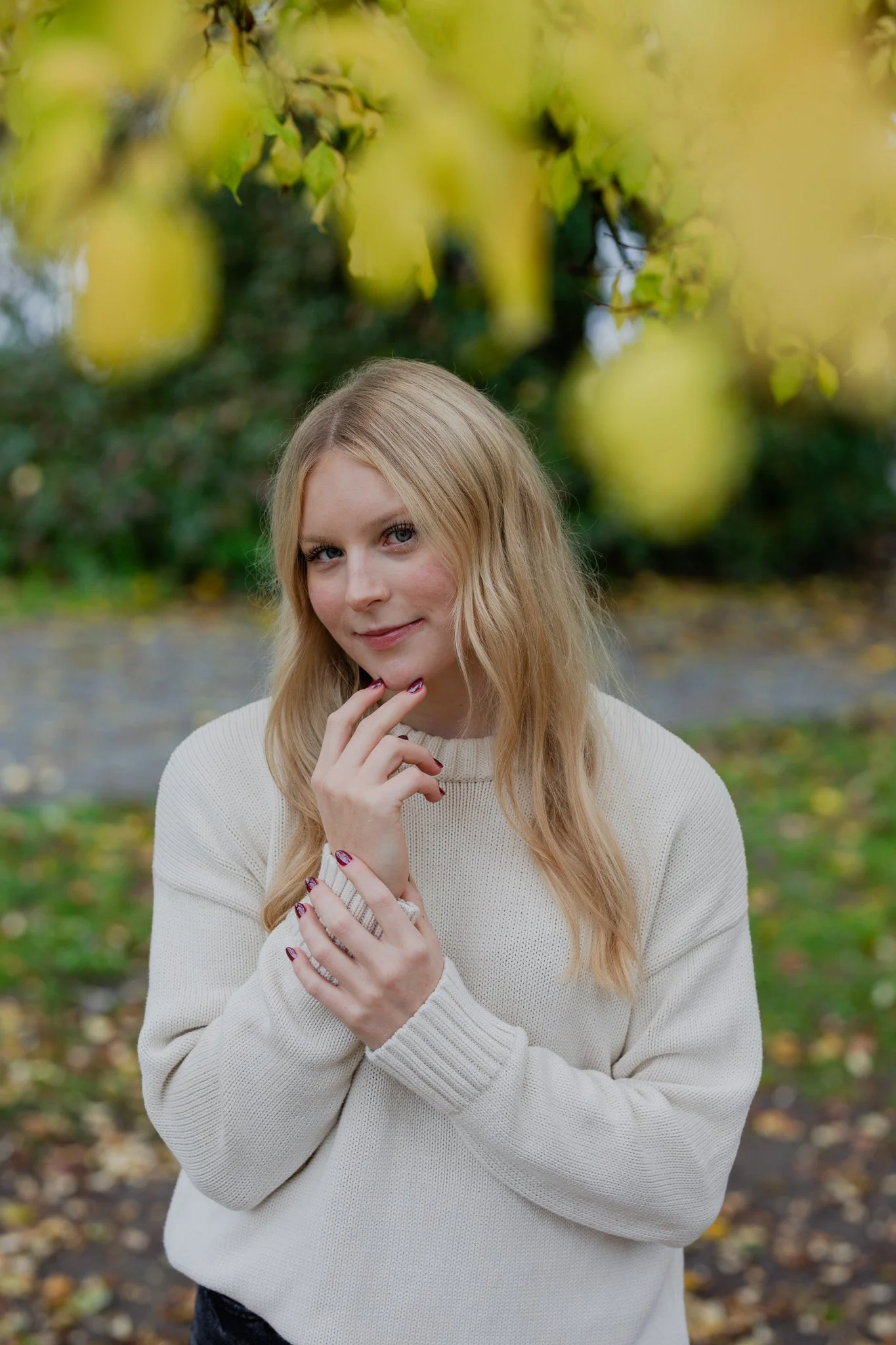 A young woman with long blonde hair and fair skin standing outdoors among yellow and green autumn leaves, wearing a cream-colored sweater, smiling and touching her face with one hand.