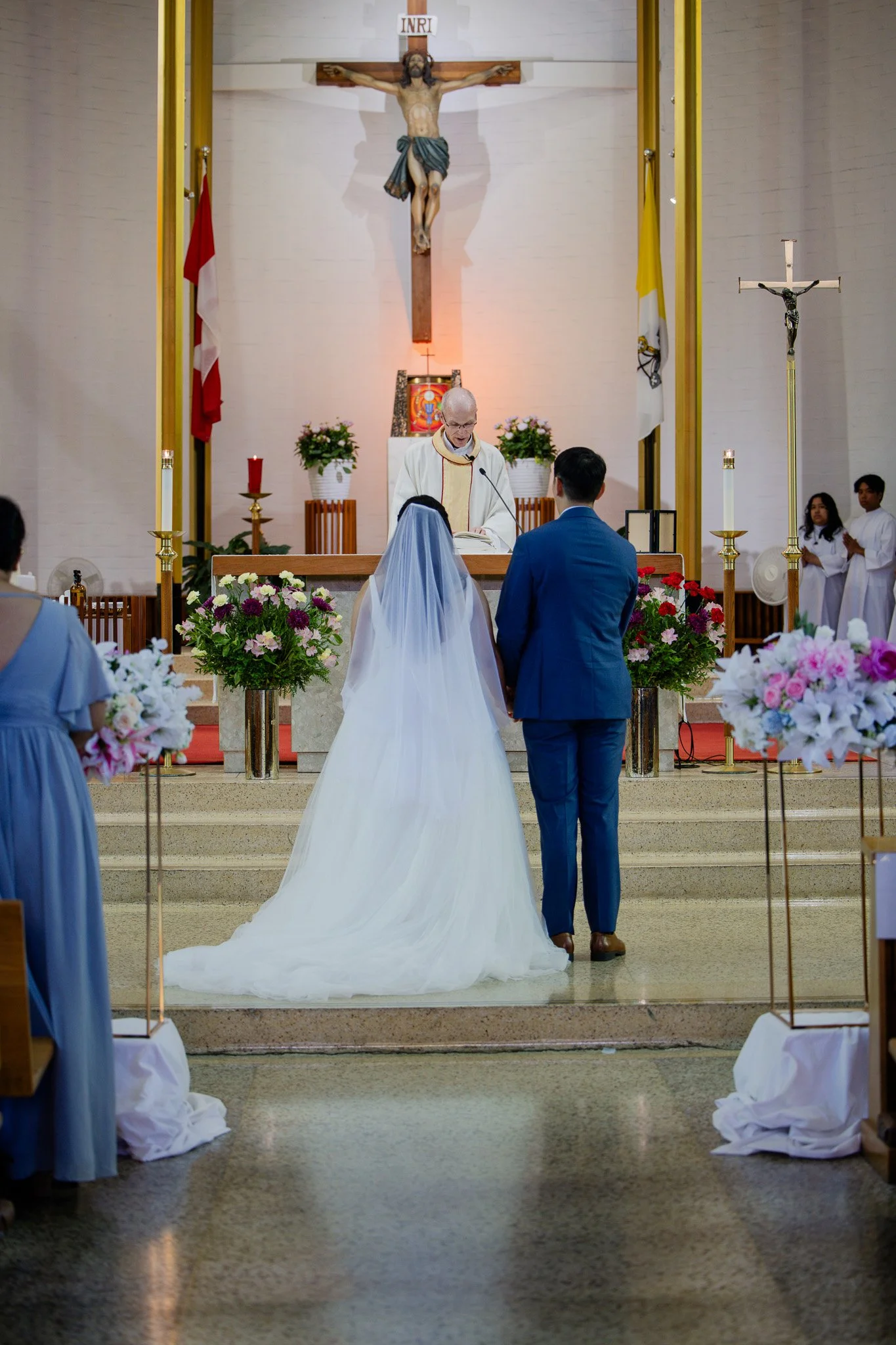A wedding ceremony inside a church with a bride and groom kneeling before a priest. The altar features large crucifixes, floral arrangements, and religious icons in the background.