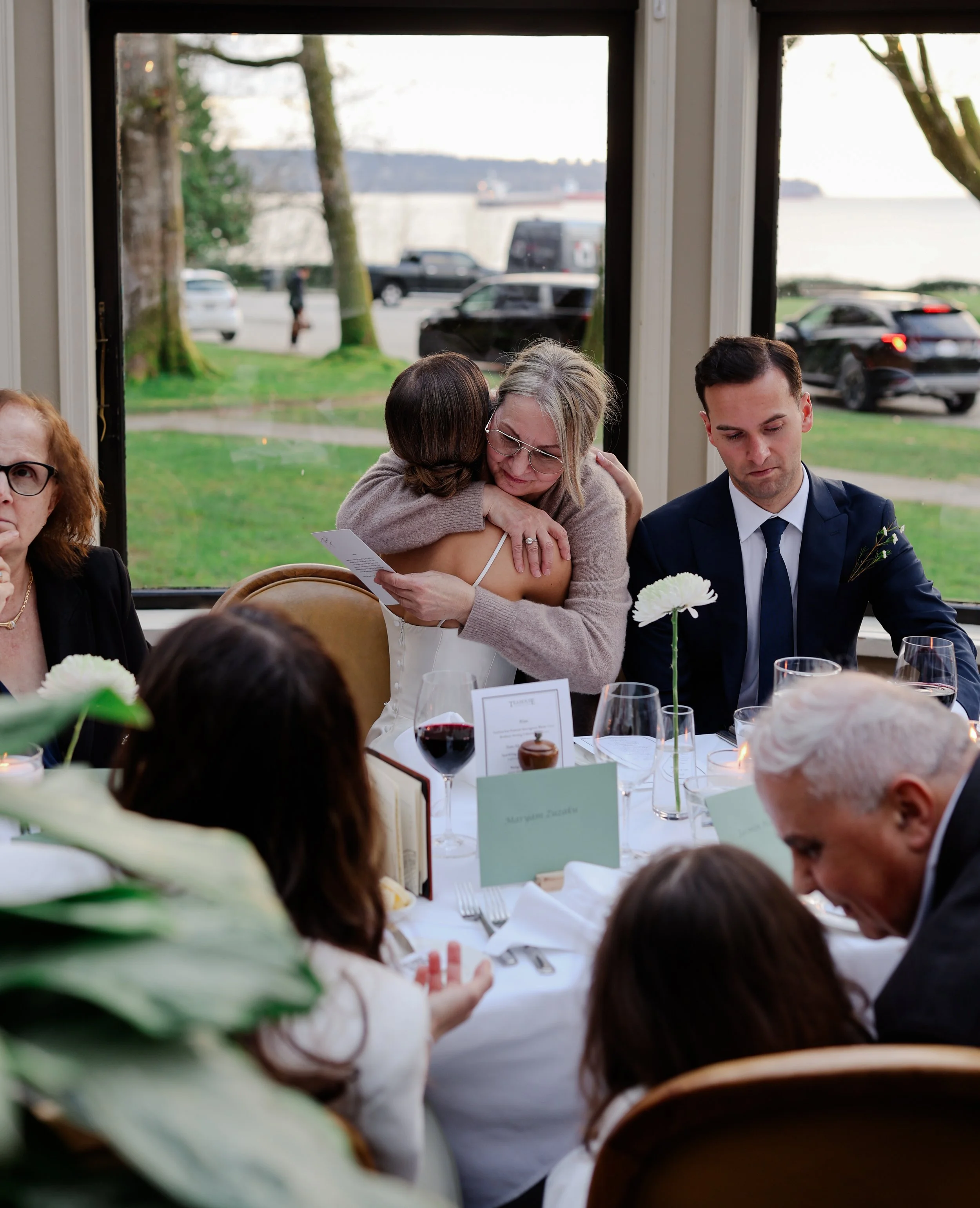 People sitting at a dining table during a celebration, with two women hugging, one wearing glasses and the other in a white dress, and another man in a suit looking down, with a window showing parked cars and trees outside.