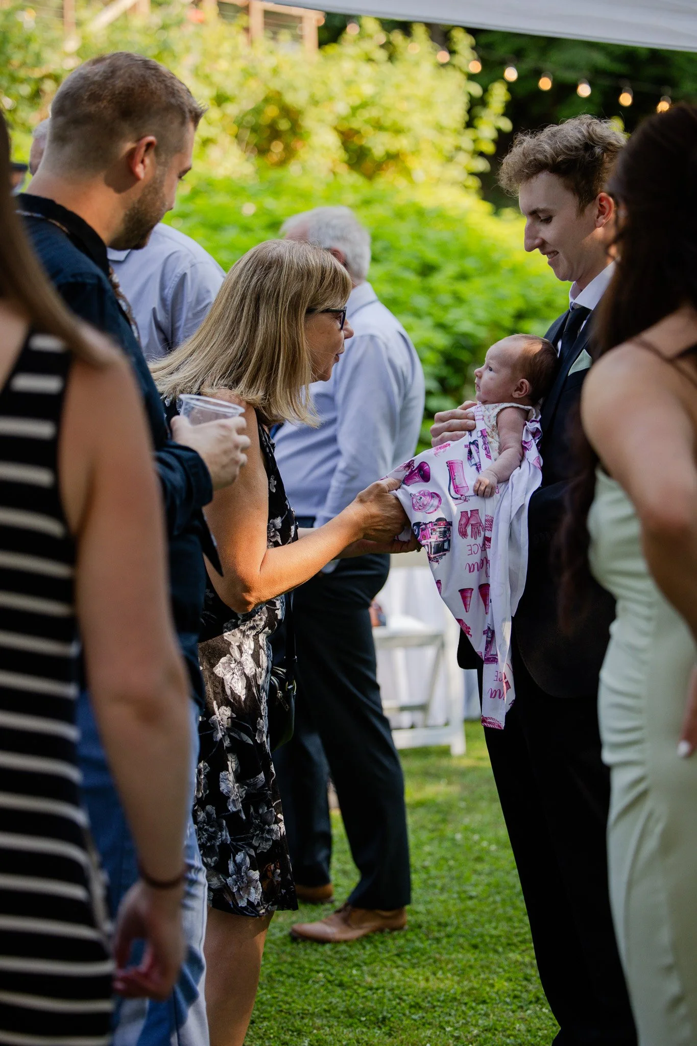 People gathered outdoors at a social event, with a woman holding a baby, wearing a dress with ice cream cone print, engaging with a man in a suit, surrounded by other individuals standing in a line on a grassy area.