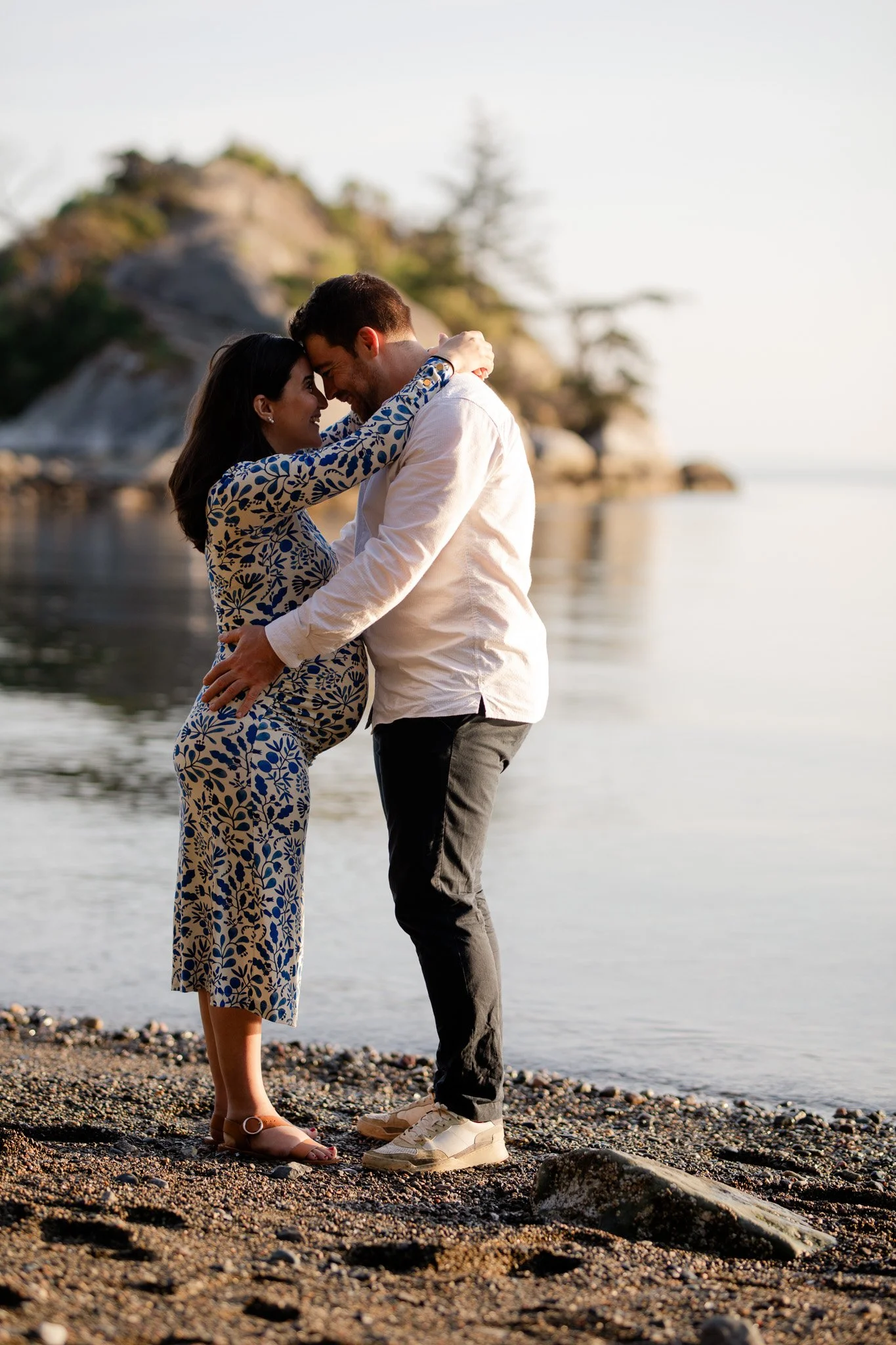 A couple embraces on a beach at sunset, with a rocky landscape and trees in the background.