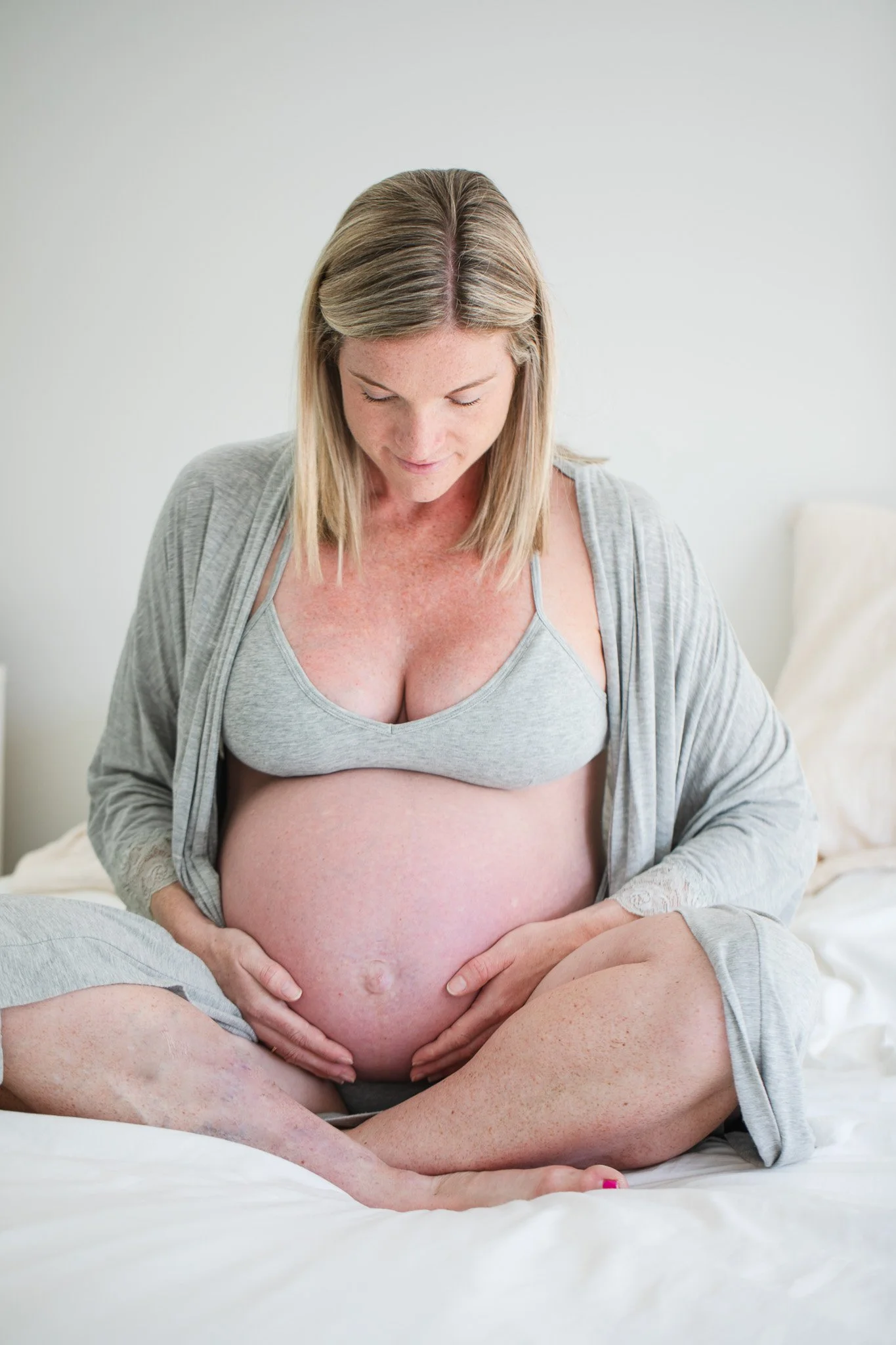 Pregnant woman sitting cross-legged on bed, looking down, wearing a gray tank top and light cardigan.