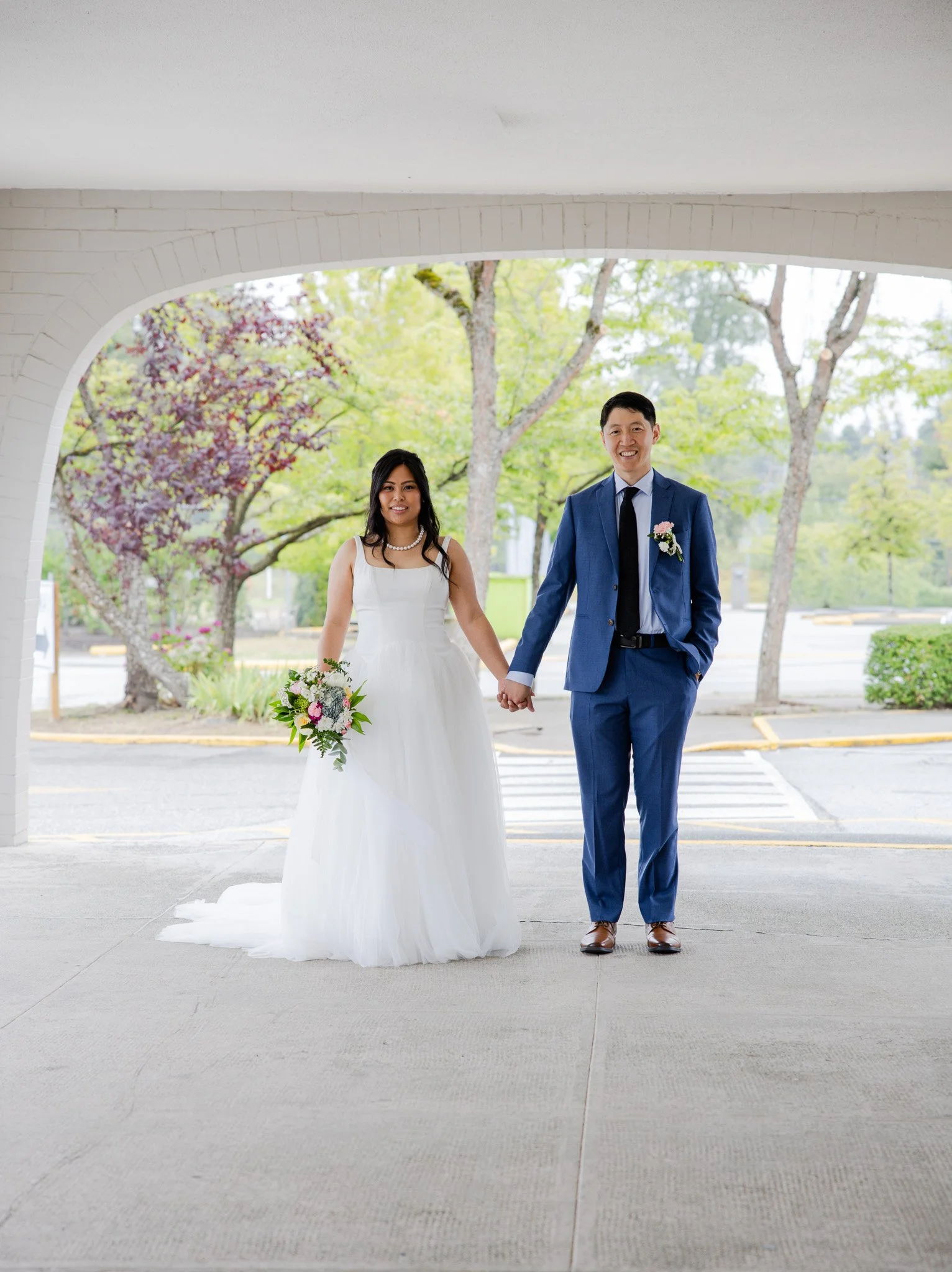 A bride and groom holding hands and smiling, standing under a white archway outdoors with trees in the background.