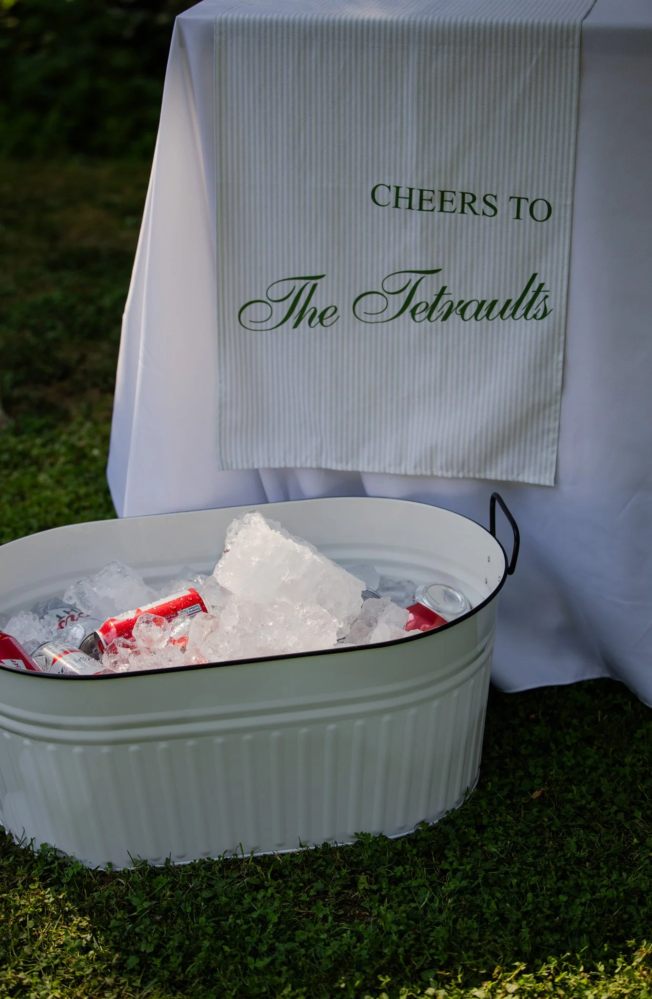 A white table with a white cloth is set outdoors, with a sign that reads 'Cheers to The Tetraults' hanging behind it. In front of the table, there is a silver container filled with ice and cans of soda.