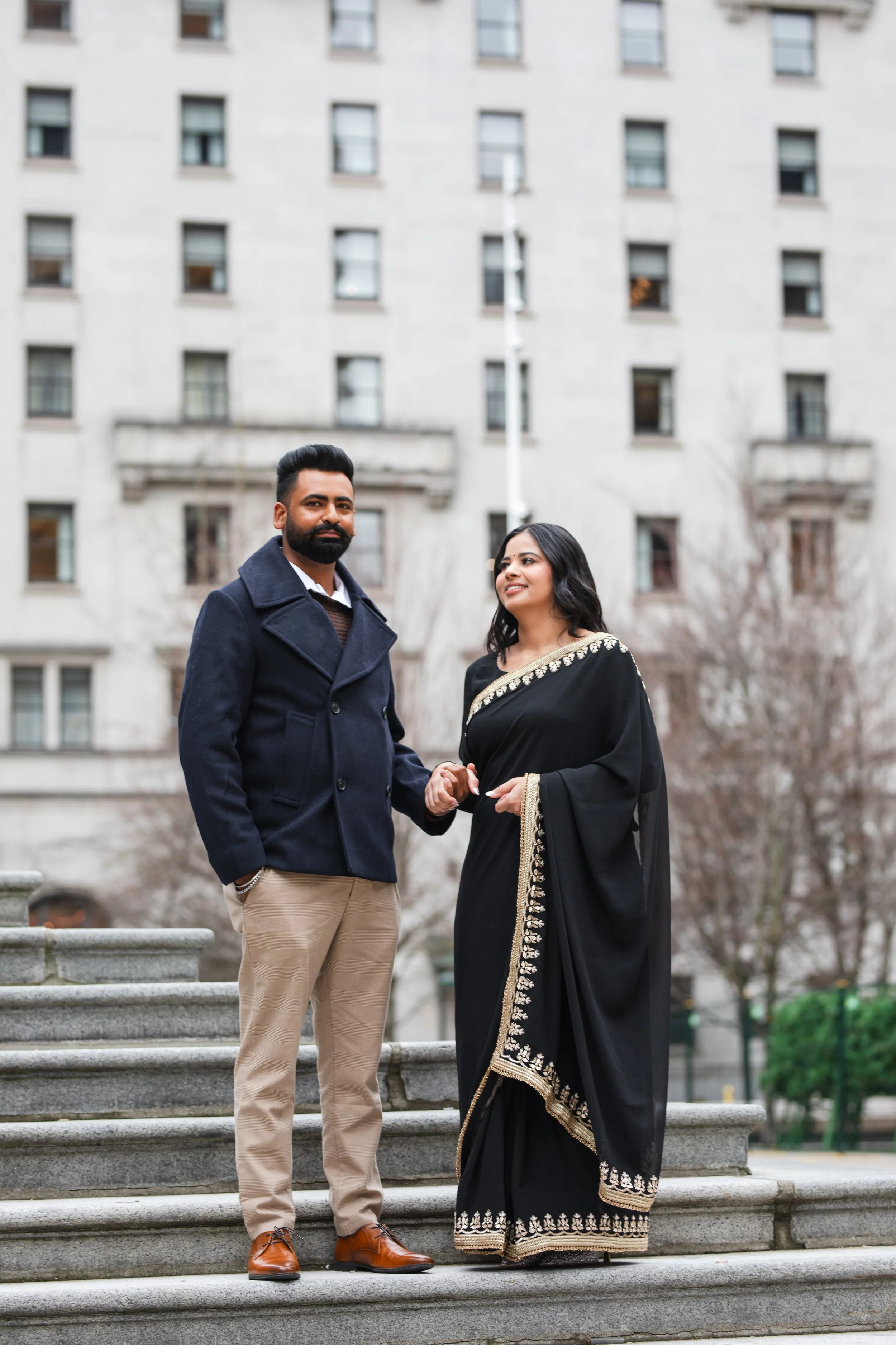 A man and a woman holding hands on a stone staircase outdoors, with a large building in the background. The man has a beard, is wearing a navy coat, beige pants, and brown shoes. The woman is wearing a black sari with gold embroidery.