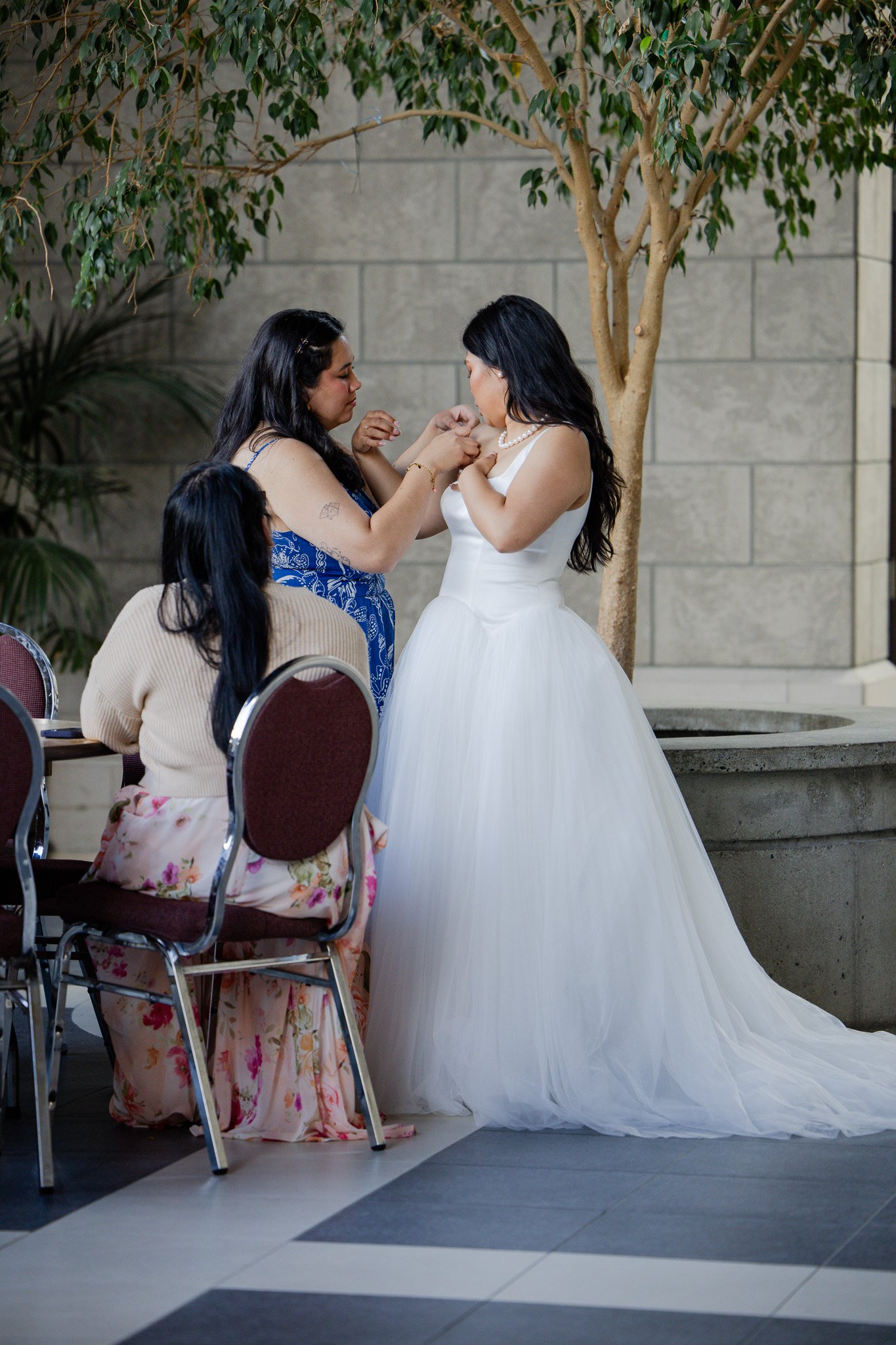 A bride in a white wedding dress is receiving a corsage from a woman in a blue dress during a wedding ceremony indoors. Other guests are seated nearby, with one woman in a floral skirt and beige sweater.