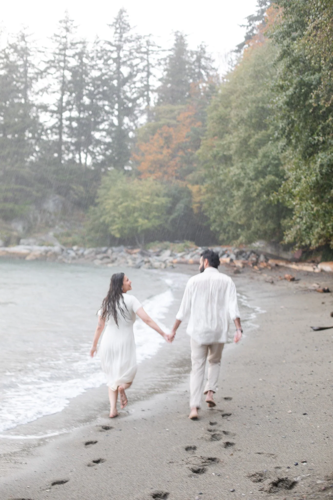 A couple holding hands walking barefoot on a sandy beach during a light rain, with trees and rocks in the background.