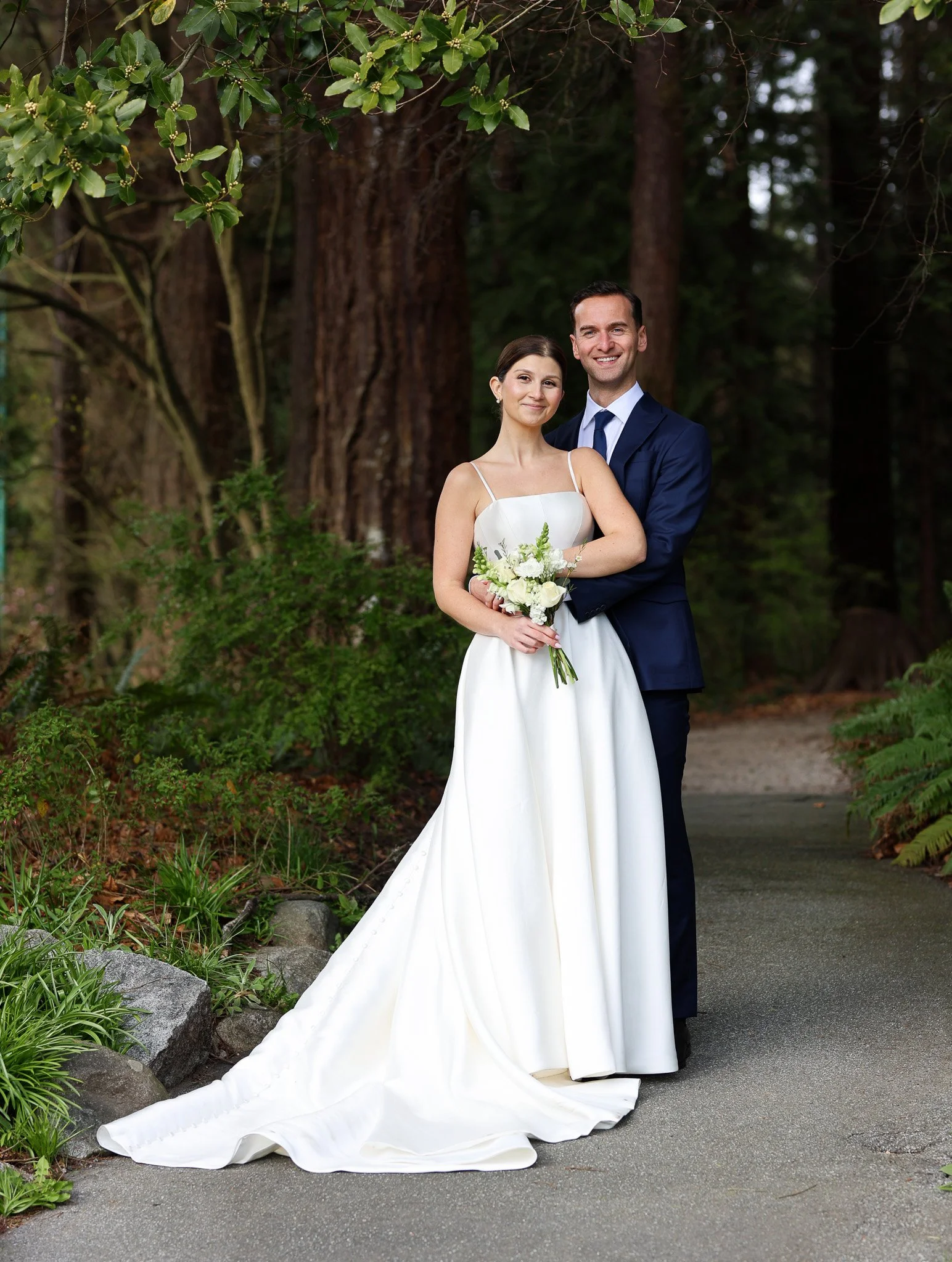 A bride and groom standing outdoors surrounded by trees and greenery. The bride is in a white wedding dress holding a bouquet, and the groom is in a navy blue suit with a white shirt and tie, both smiling.