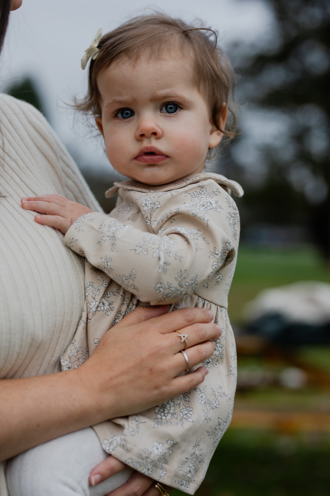 A young girl with blue eyes and light brown hair, wearing a beige floral dress, is being held by an adult outdoors. The child looks directly at the camera with a serious expression, a small bow in her hair, and her hand resting on the adult's arm.