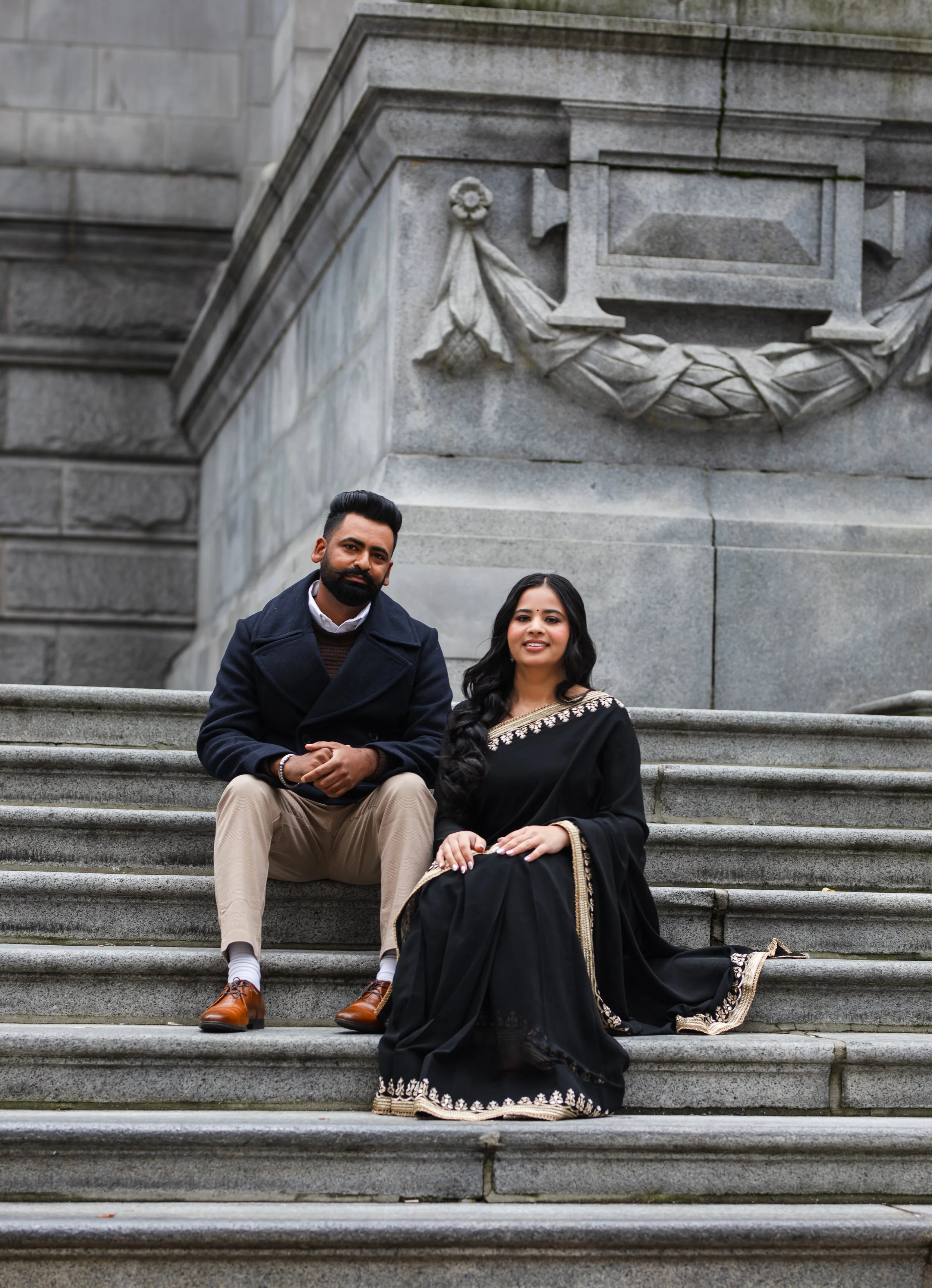 A man and woman sitting on stone steps in front of a large stone monument with decorative carvings. The woman is wearing a black saree with gold embroidery, and the man is dressed in a dark coat, beige pants, and brown shoes.